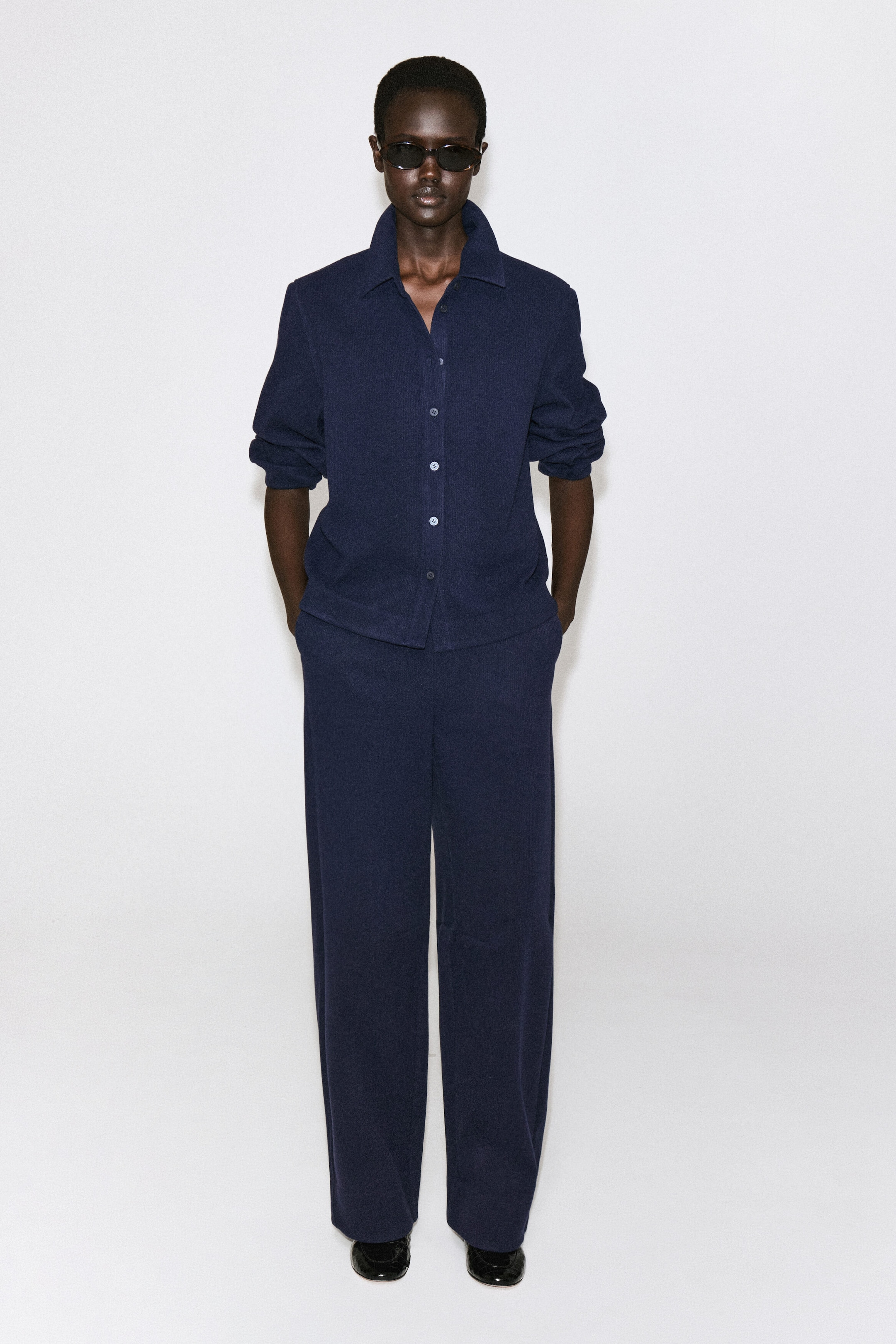 This navy blue corduroy jersey shirt is shown from the front, featuring a relaxed fit, a classic collar, and a front button closure. The long sleeves are worn pushed up to the elbows, and the shirt is paired with matching trousers. This navy blue corduroy jersey shirt is shown from the front, featuring a relaxed fit, a classic collar, and a front button closure. The long sleeves are worn pushed up to the elbows, and the shirt is paired with matching trousers.