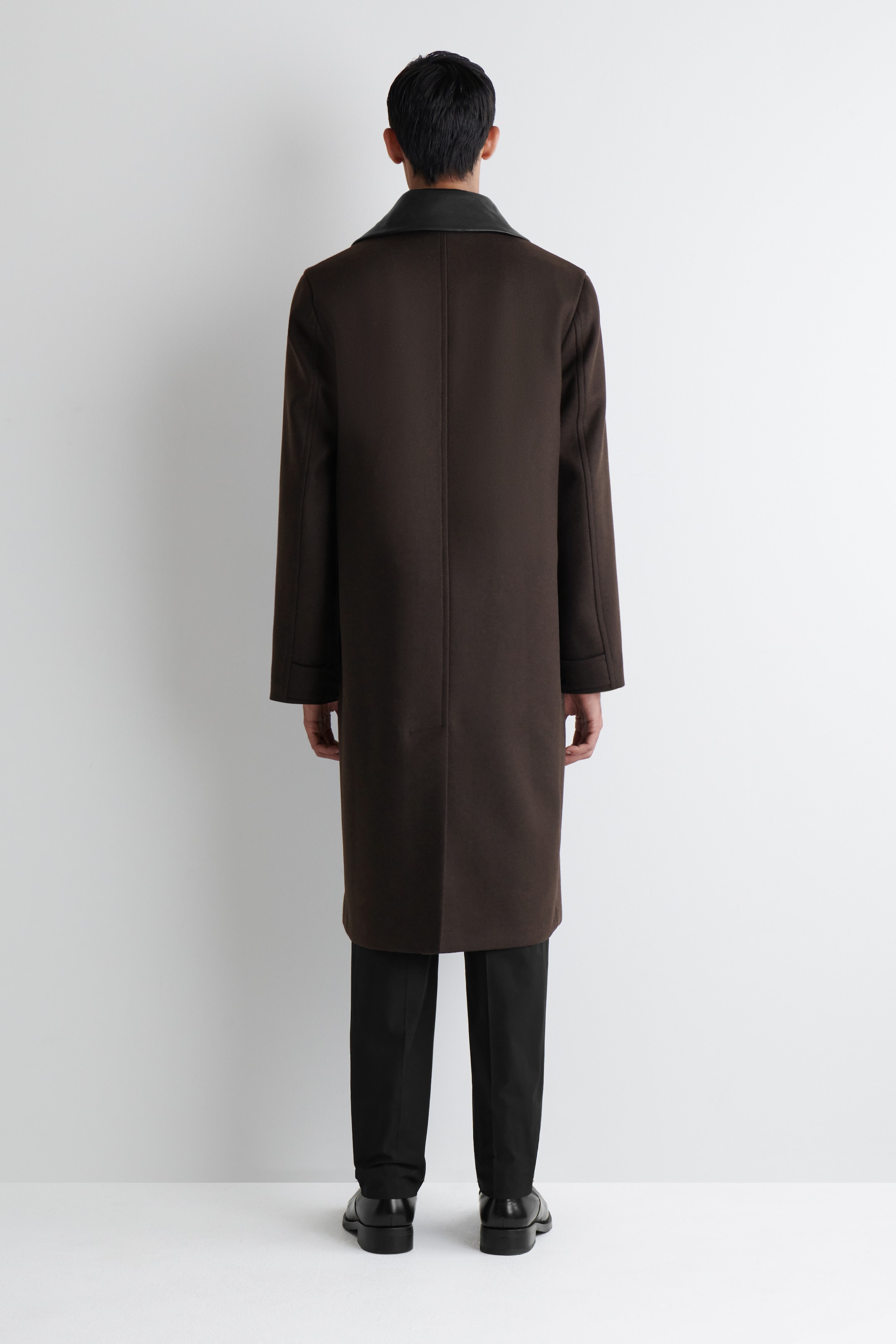 Visualizza immagine più grande: Un cappotto di lana marrone scuro viene mostrato dal RETRO, caratterizzato da una vestibilità rilassata e una lunghezza che si estende oltre le ginocchia. È progettato con un colletto in pelle scura a contrasto e una cucitura centrale sul retro con uno spacco.