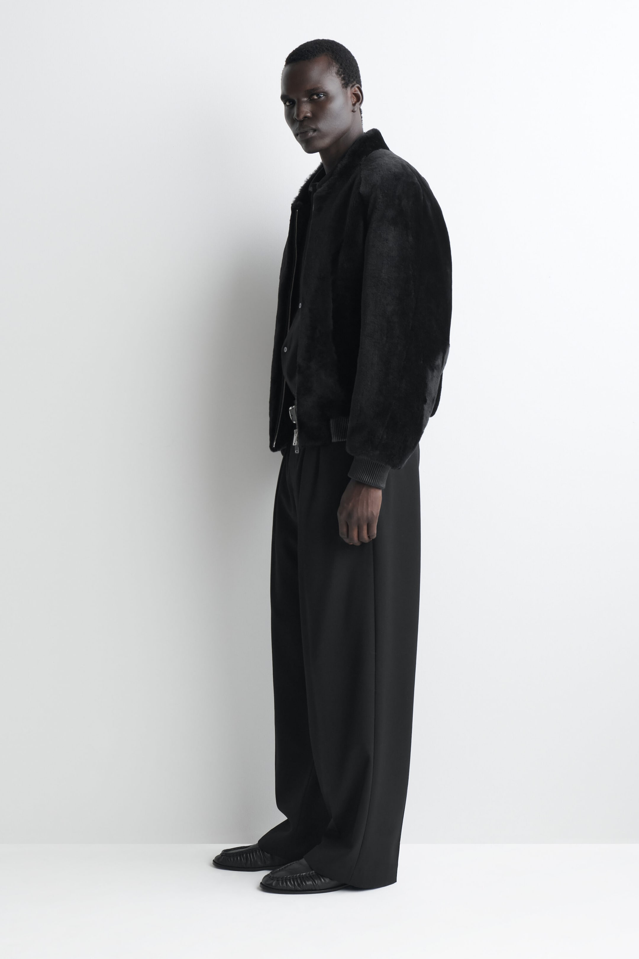 A black padded shearling bomber jacket is facing left, showcasing its plush, textured exterior and relaxed, voluminous silhouette. The jacket features ribbed knit cuffs and a matching ribbed hem and is layered over a dark shirt and wide-leg trousers.