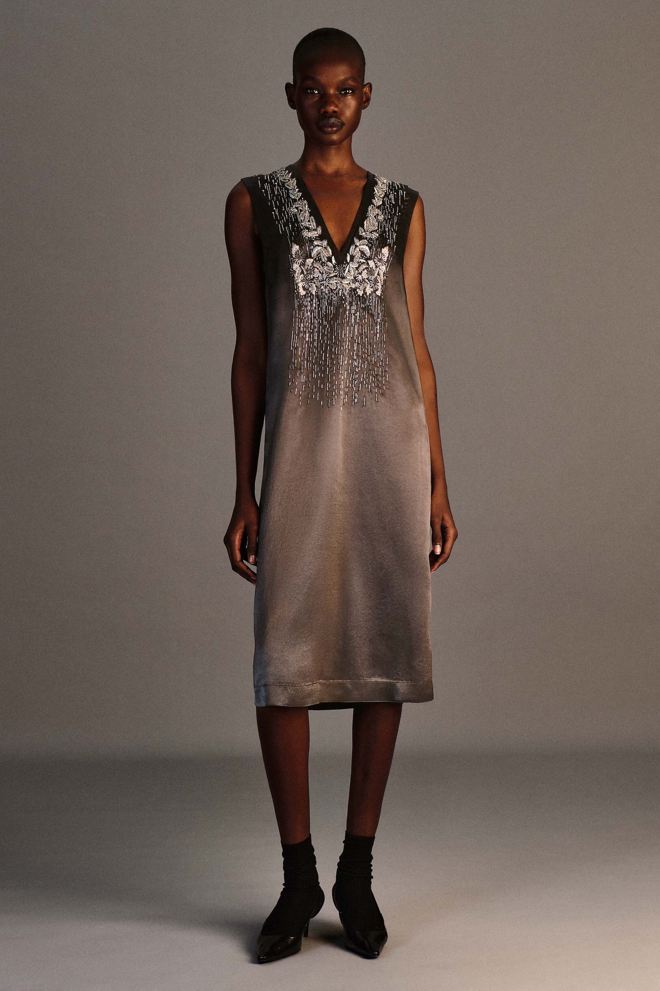 A beaded satin V-neck midi dress is showcased in a front view, featuring a sleeveless, straight-cut silhouette in gradient gray-to-silver satin fabric. The dress features intricate silver floral embroidery and beadwork around the V-neckline, cascading down the chest in vertical lines. A beaded satin V-neck midi dress is showcased in a front view, featuring a sleeveless, straight-cut silhouette in gradient gray-to-silver satin fabric. The dress features intricate silver floral embroidery and beadwork around the V-neckline, cascading down the chest in vertical lines.