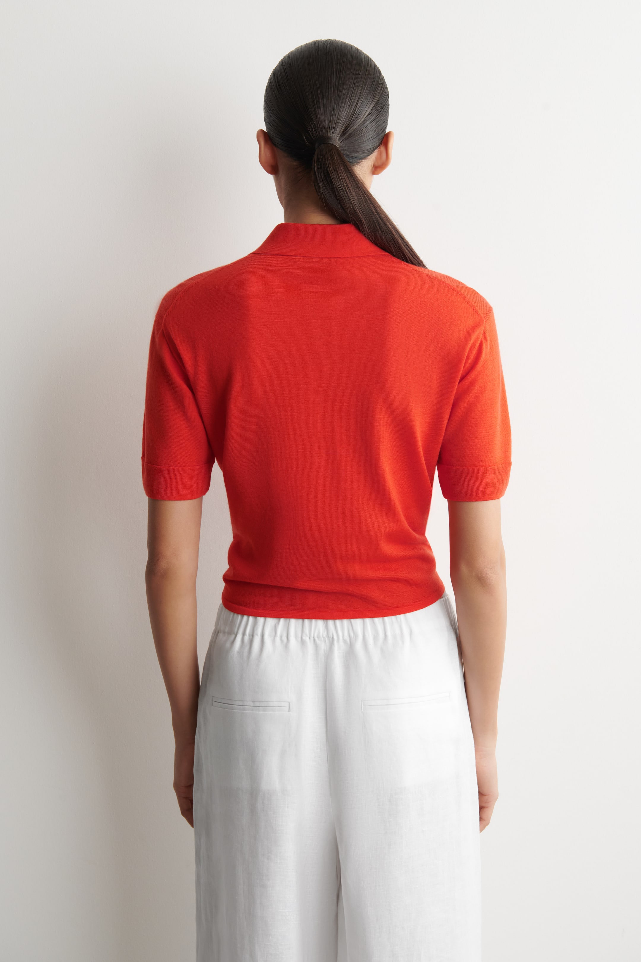 Grotere afbeelding bekijken: Een rode gebreide polo met korte mouwen wordt van de achterkant gepresenteerd, met een platte polokraag en omgeslagen korte mouwen. De zoom is ingestopt in witte broek.