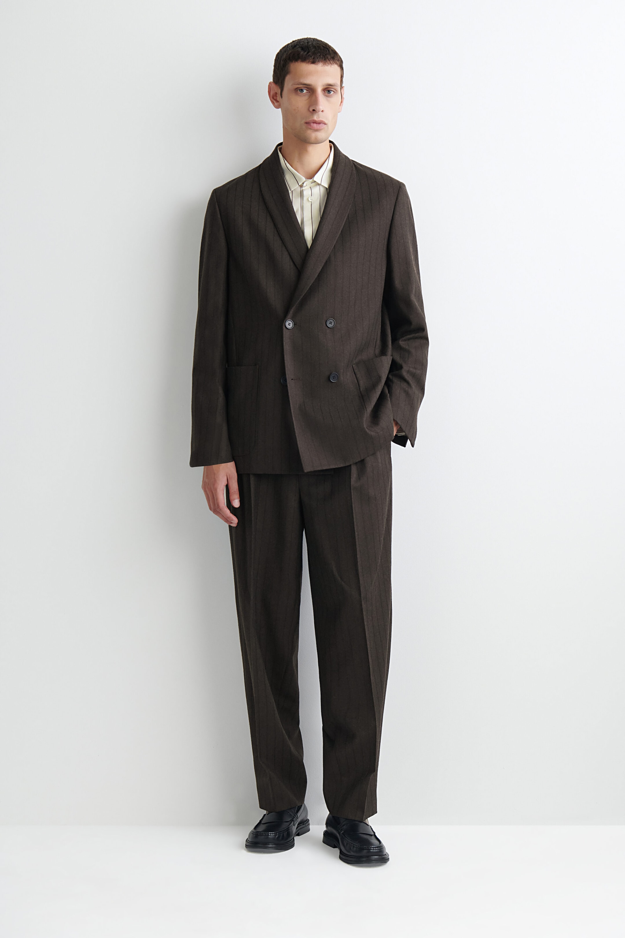 A model is wearing a dark brown, pinstriped double-breasted blazer in a relaxed fit, featuring shawl-style lapels and side pockets, shown from the front. The blazer is styled as a suit with matching wide-leg trousers, a light-colored striped collared shirt, and black leather loafers. A model is wearing a dark brown, pinstriped double-breasted blazer in a relaxed fit, featuring shawl-style lapels and side pockets, shown from the front. The blazer is styled as a suit with matching wide-leg trousers, a light-colored striped collared shirt, and black leather loafers.