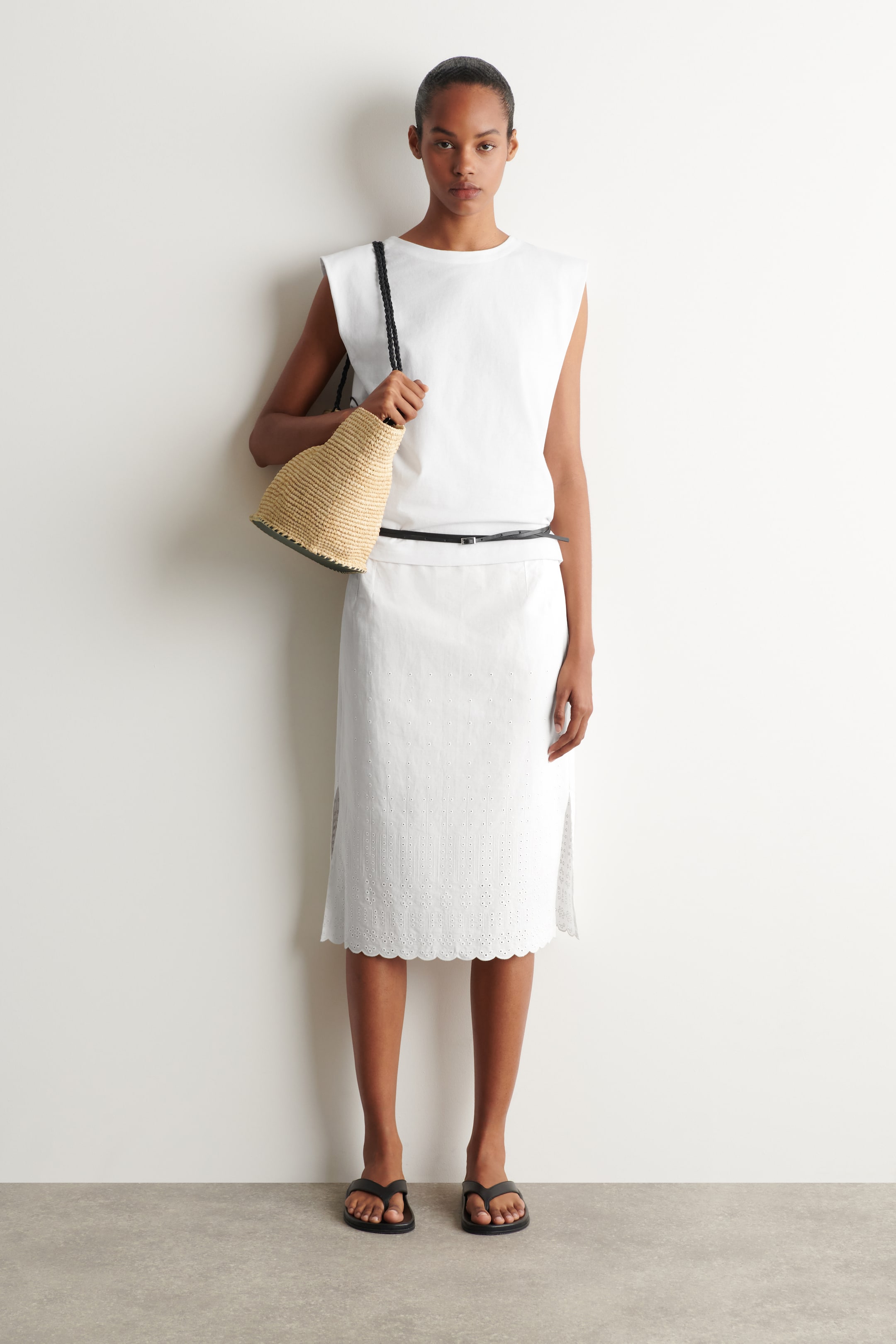 View larger image: A white sleeveless top featuring structured shoulders and a crew neck is paired with a white knee-length skirt showcasing eyelet embroidery and a scalloped hem. A slim black belt cinches the waist, while a small woven straw bucket bag with a black braided strap is carried, and black thong sandals complete the ensemble.