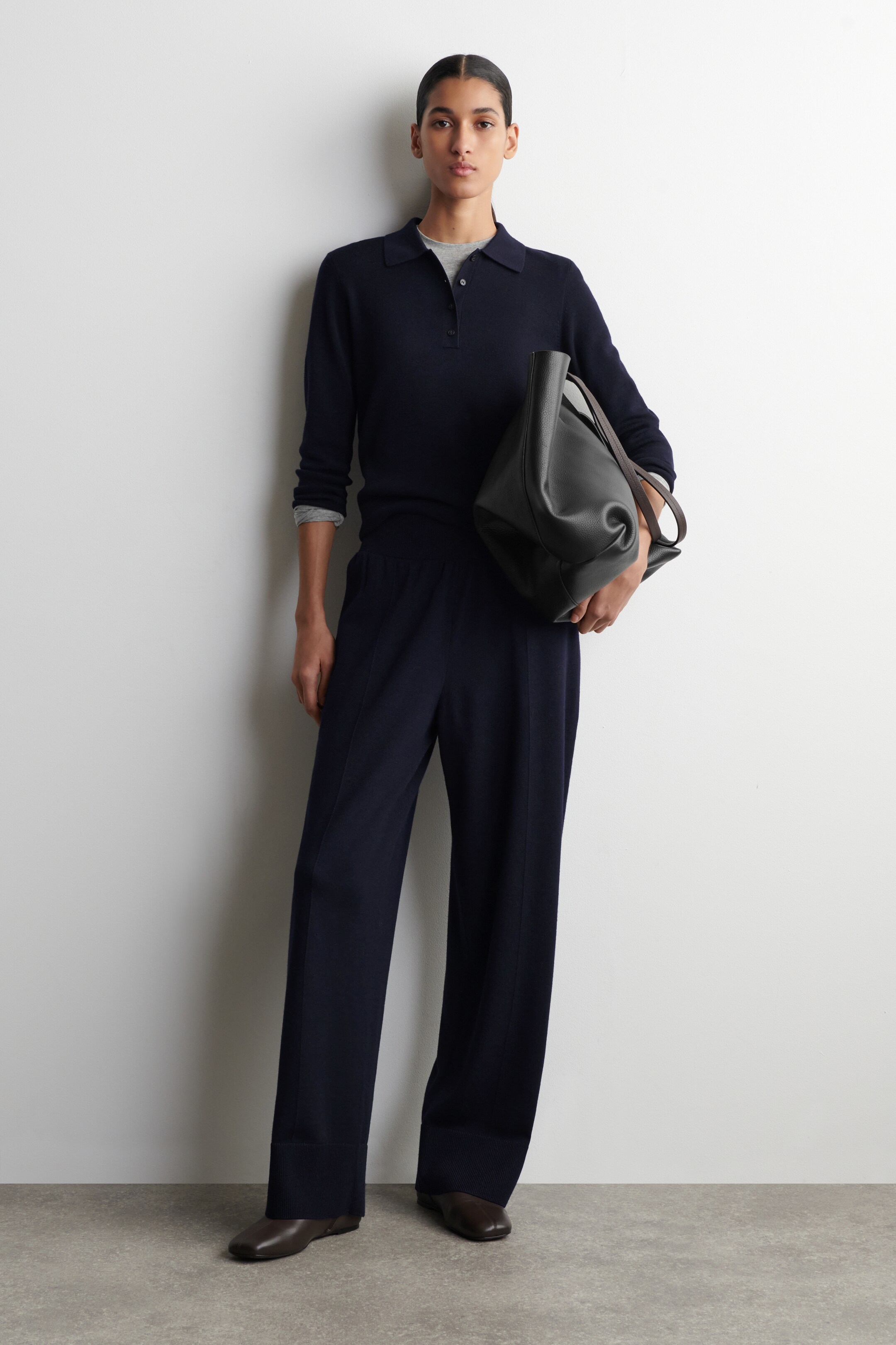 View larger image: A complete outfit is showcased from a full front angle, featuring a navy long-sleeved polo jumper with a collar and a three-button placket, layered over a grey crew neck t-shirt and paired with navy wide-leg trousers displaying a stitched front seam on each leg. A black leather tote bag with a brown shoulder strap is also visible, along with brown square-toe flat shoes.