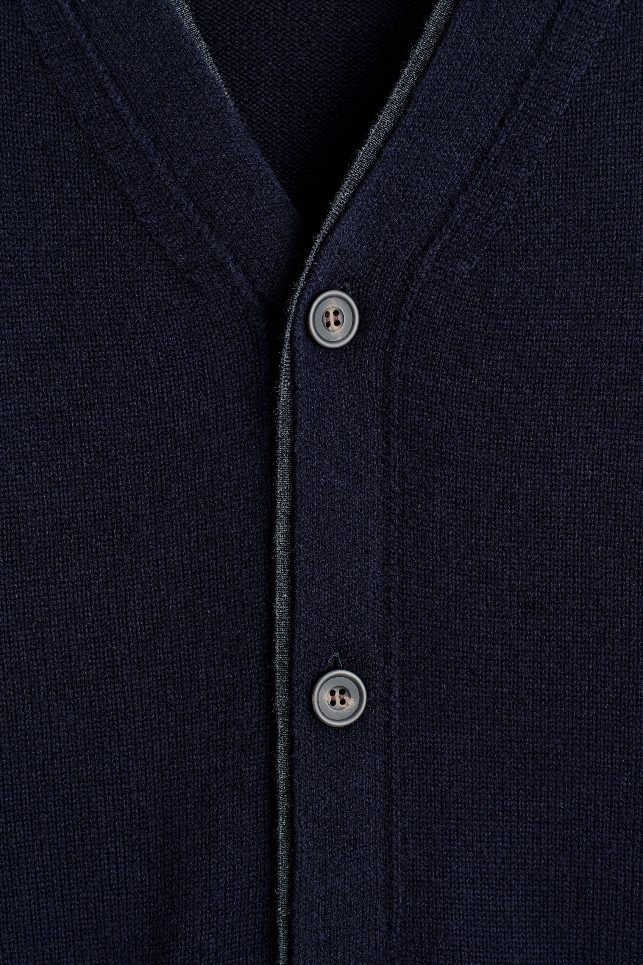 Grösseres Bild ansehen: Eine marineblaue Strickjacke wird von vorne präsentiert, mit einem V-Ausschnitt und einer Knopfleiste. Die Leiste ist mit zwei sichtbaren Vierlochknöpfen und einem kontrastierenden grauen Besatz versehen.
