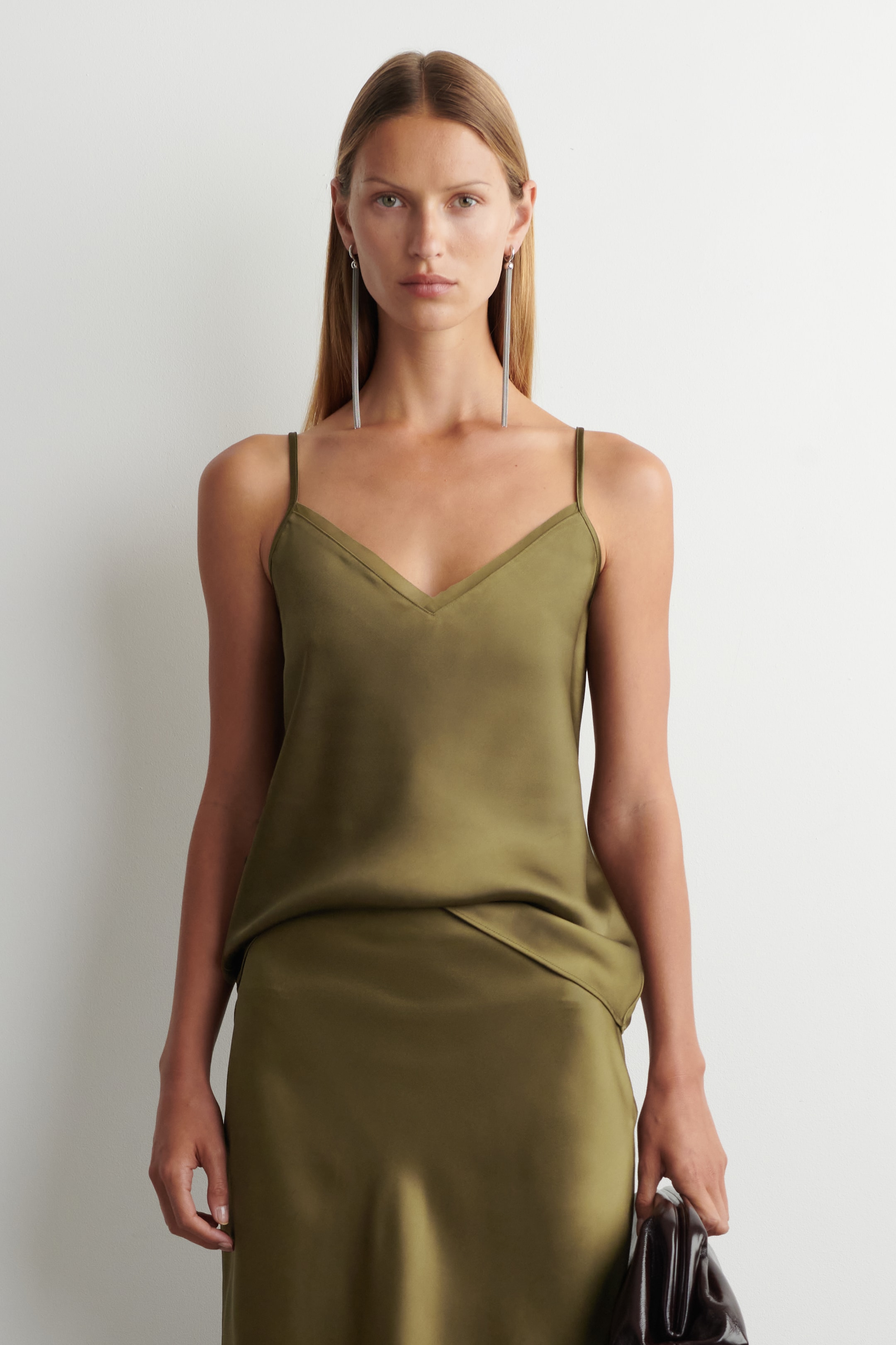 Agrandir l'image: Un haut en satin vert olive avec de fines bretelles spaghetti et un décolleté en V, subtilement rentré dans une jupe midi en satin vert olive assortie. Le tissu tombe doucement, et l'ensemble présente une vue coordonnée de face.