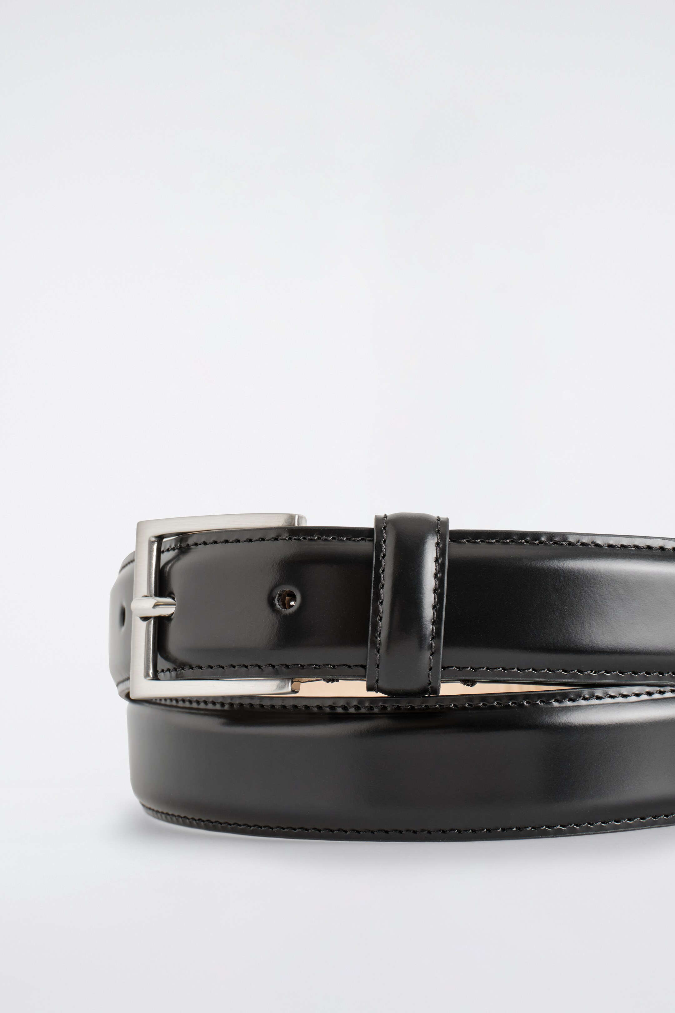 View larger image: A black leather belt is curving across the bottom of the frame, showcasing a smooth, glossy finish. A rectangular, brushed silver-tone buckle is securing the strap, and visible stitching is detailing the edges of the leather.