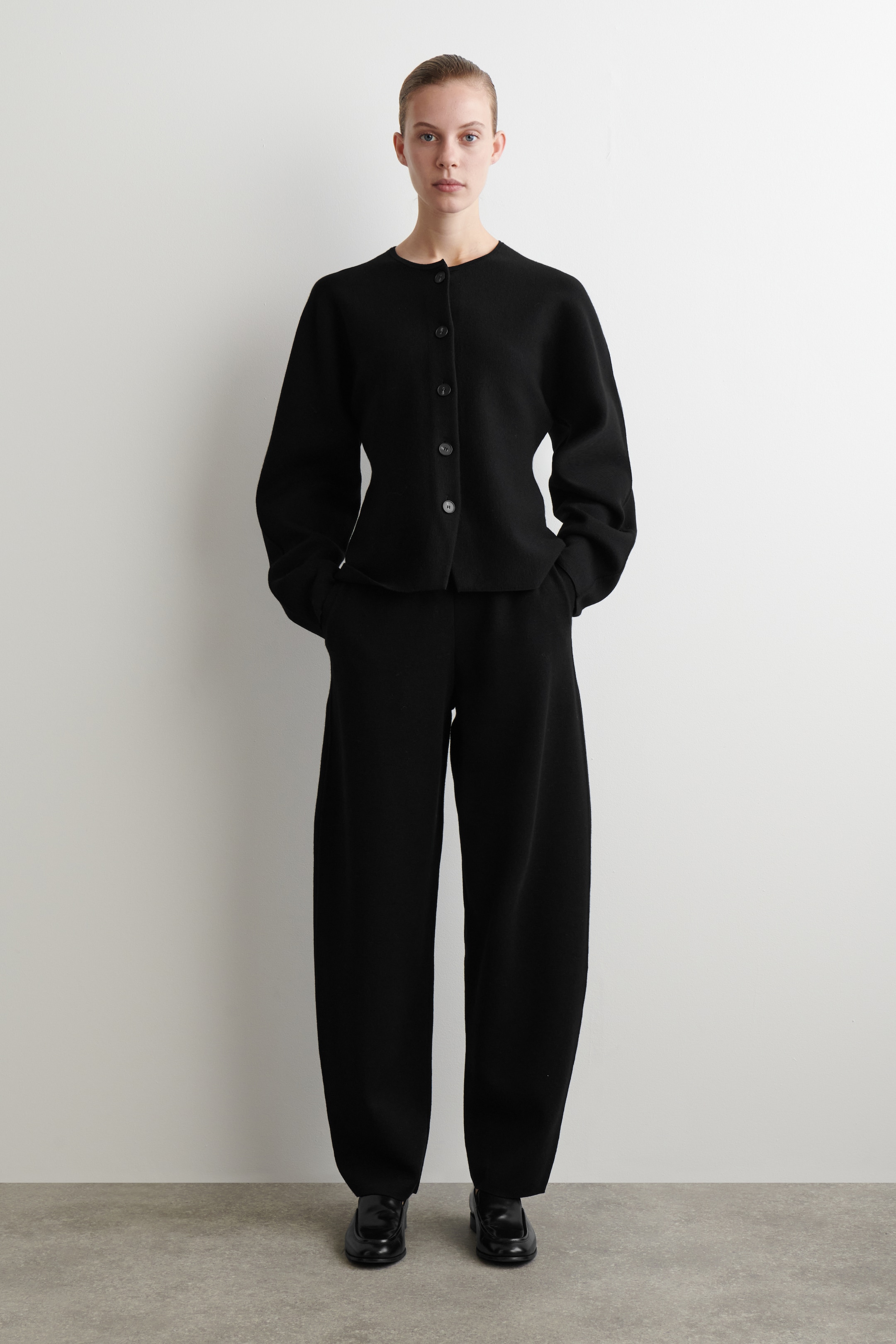 Visualizza immagine più grande: Pantaloni neri a maglia con gamba a barile, caratterizzati da una silhouette rilassata e scultorea, sono mostrati dalla parte anteriore. Sono abbinati a un cardigan nero a maglia coordinato con scollo rotondo e chiusura frontale con bottoni.
