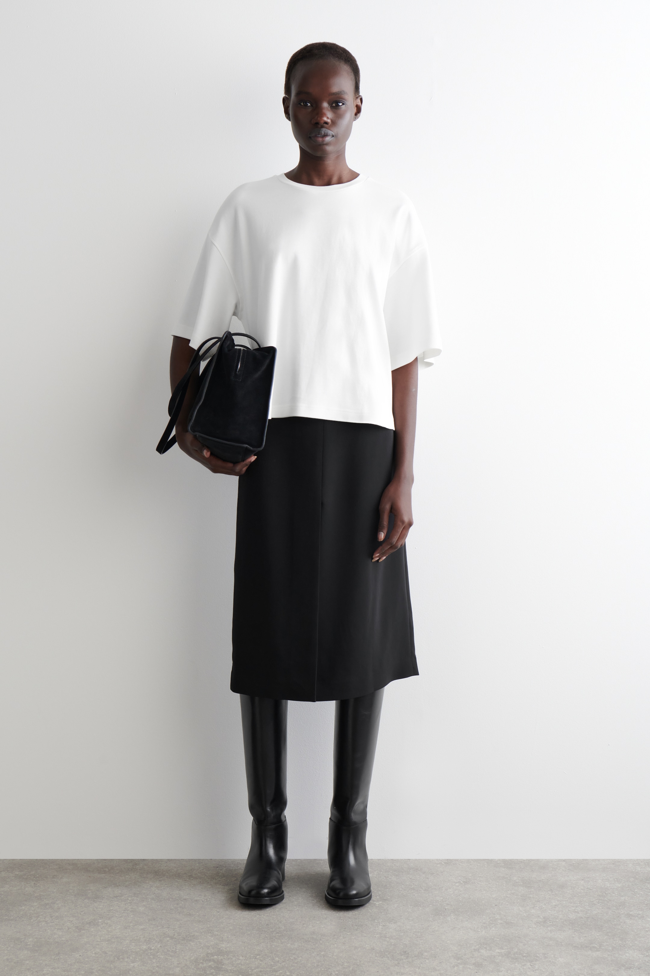 Visa större bild: En framåtvänd modell visar upp en minimalistisk ensemble, iklädd en vit, löst sittande topp med vida, korta ärmar och en rund halsringning. Hon kombinerar den med en rak, svart midikjol och höga svarta stövlar, medan hon håller i en strukturerad svart tote-väska med topphandtag.