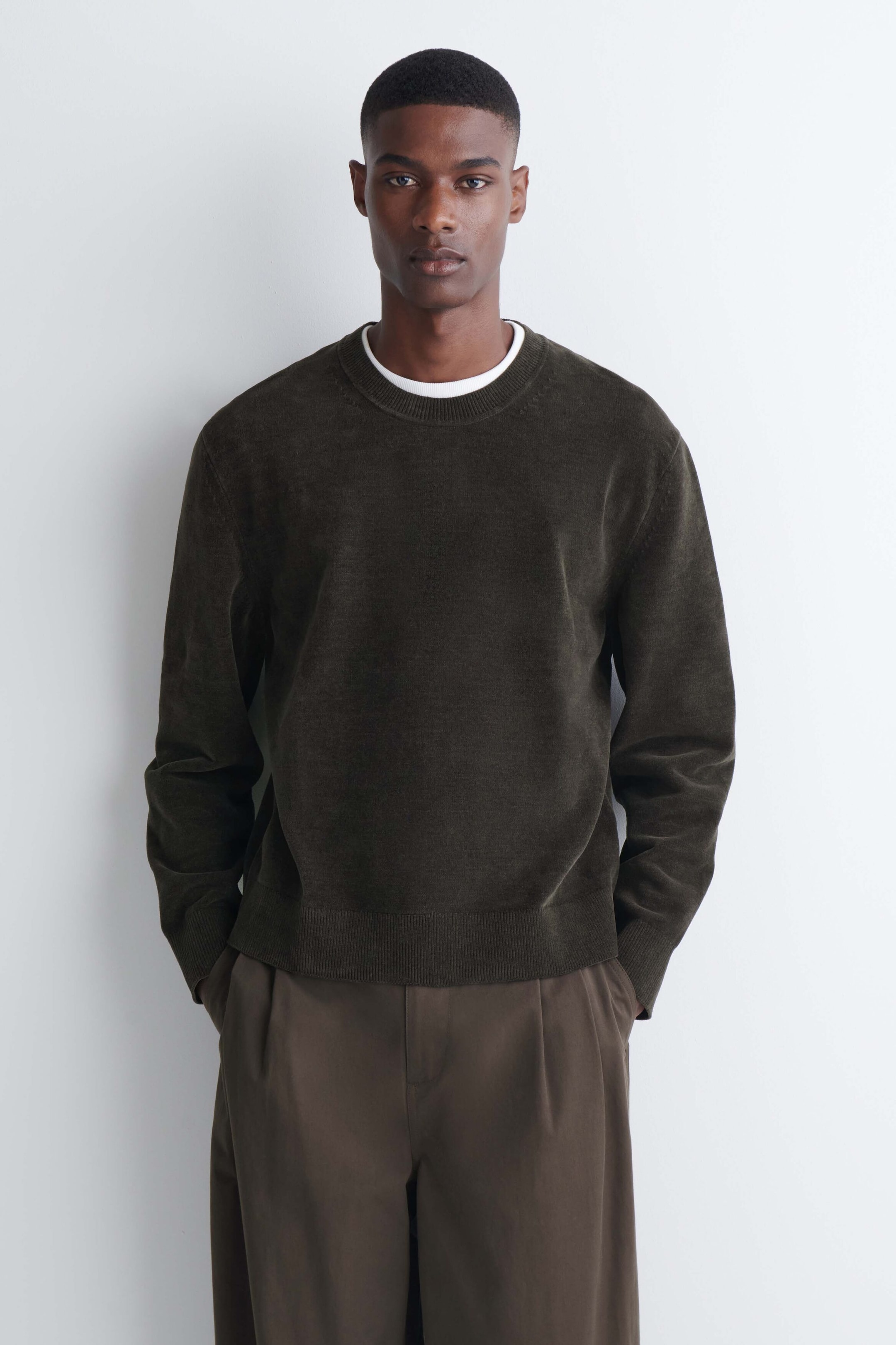 The model is wearing a dark olive green cotton chenille crew neck sweater layered over a white T shirt. The sweater features a soft, textured finish with ribbed collar, cuffs, and hem, and is paired with brown trousers. The model is wearing a dark olive green cotton chenille crew neck sweater layered over a white T shirt. The sweater features a soft, textured finish with ribbed collar, cuffs, and hem, and is paired with brown trousers.