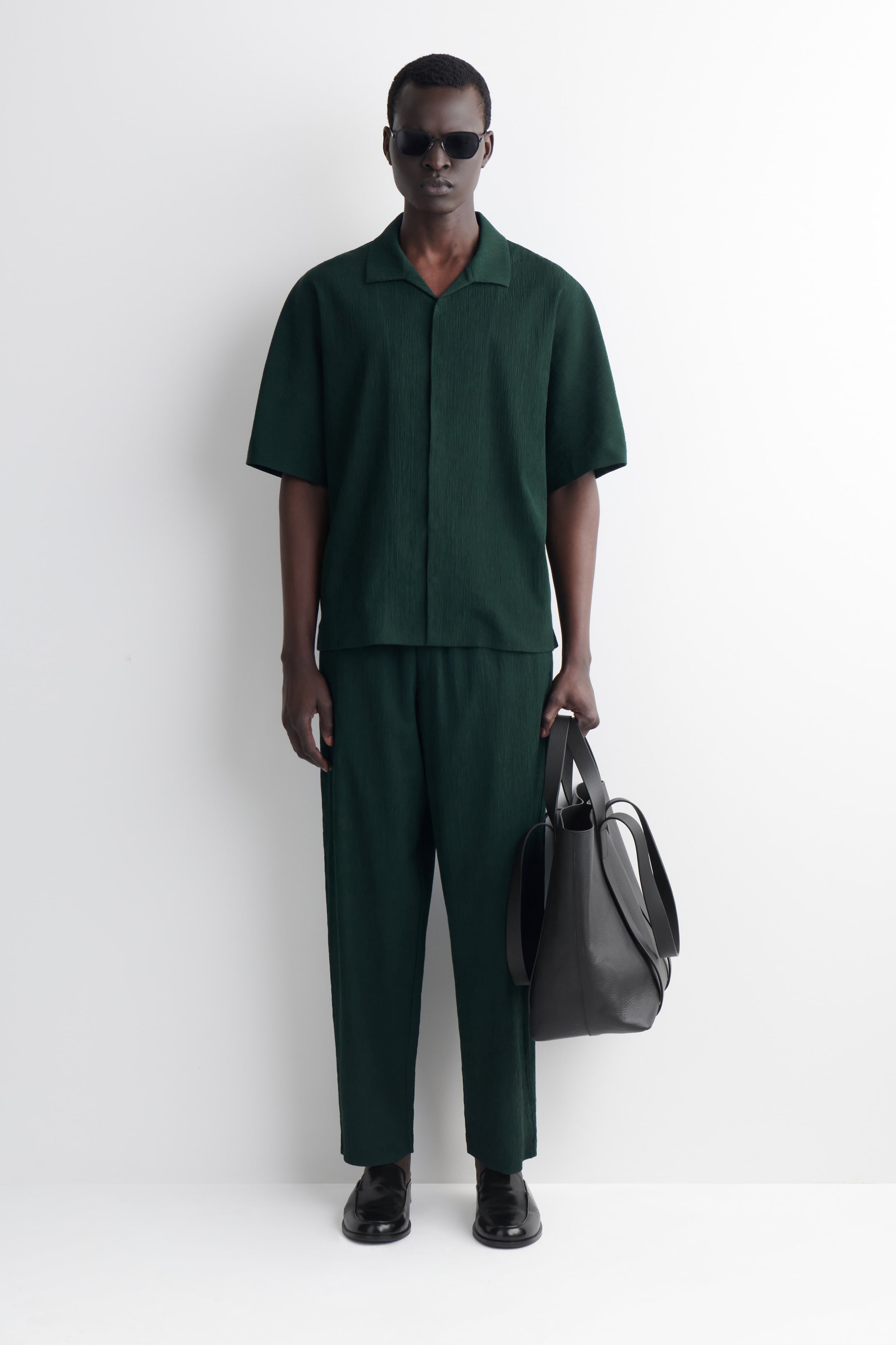Ver imagem maior: Um conjunto coordenado verde escuro, composto por uma camisa de manga curta com gola aberta e calças descontraídas, é apresentado de frente, ambas as peças com um tecido subtilmente texturizado.