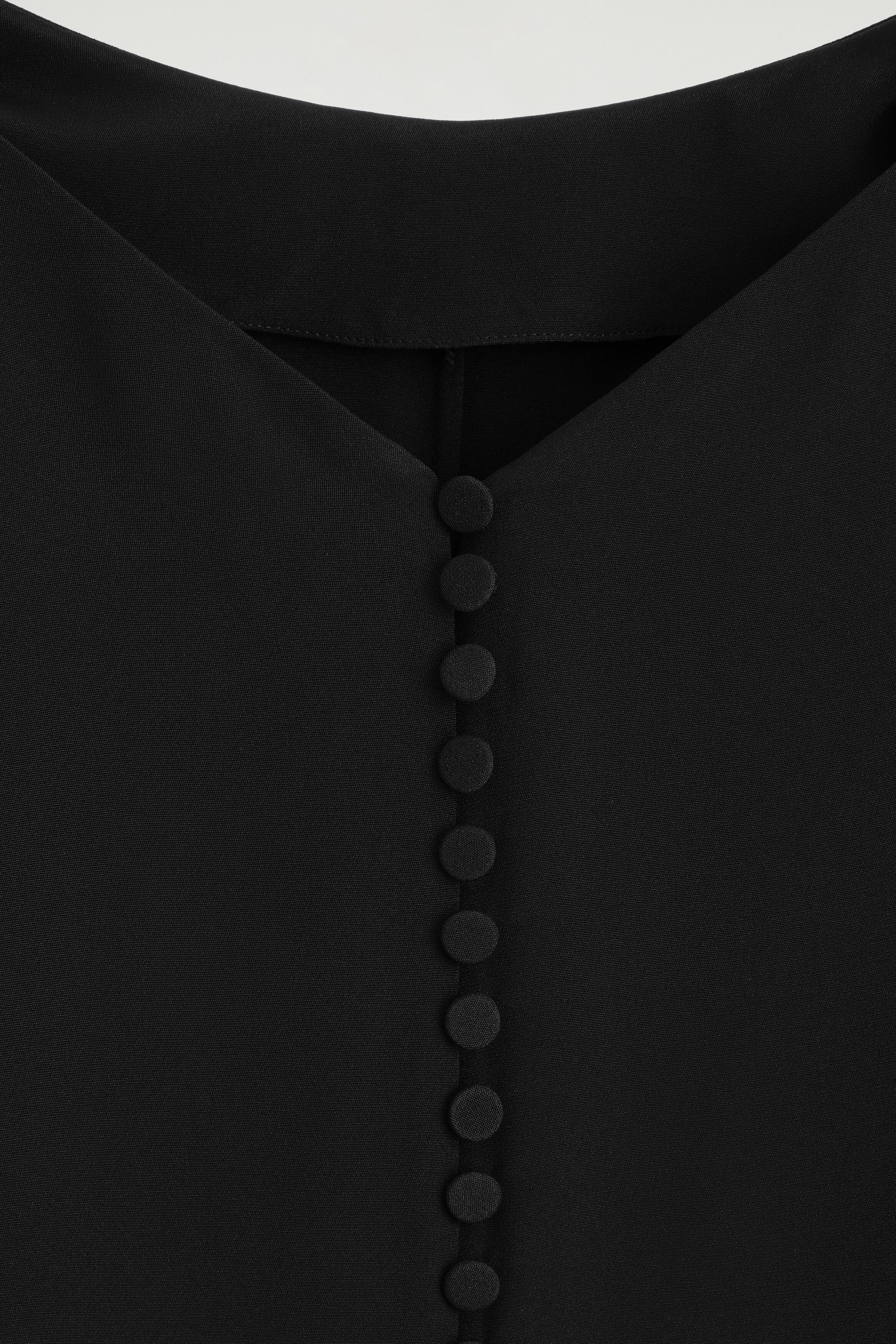A close-up view highlights the back of a black crepe maxi dress, showcasing a V-shaped neckline and a vertical row of fabric-covered buttons fastening down the centre.