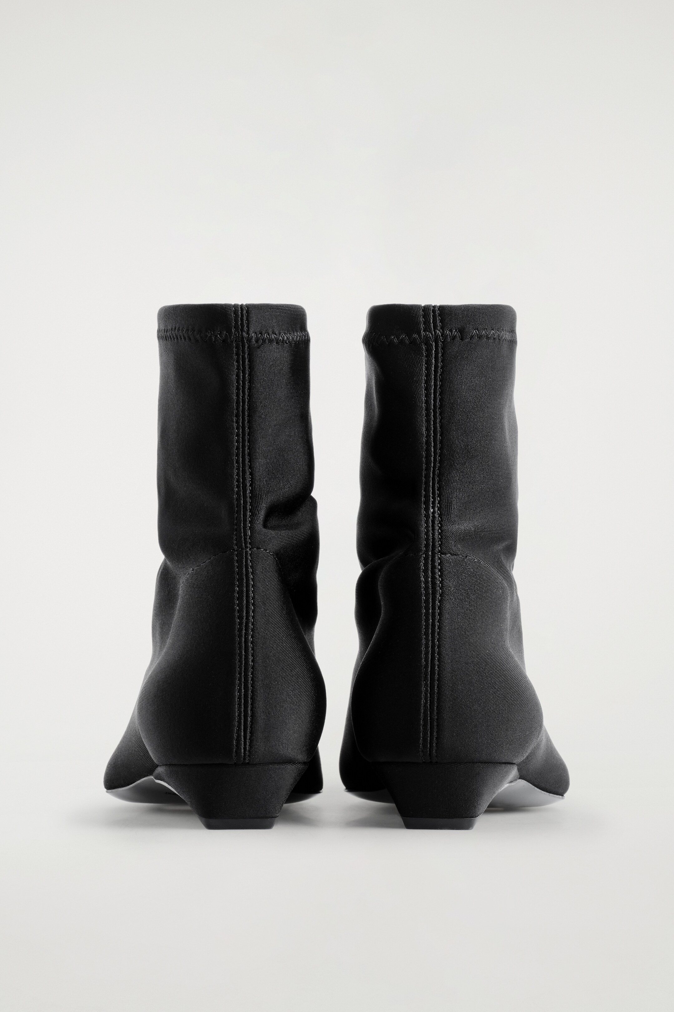 Größeres Bild ansehen: Ein Paar schwarze Sock Boots wird von hinten präsentiert, mit einem schlanken, knöchellangen Schaft aus Scuba-Material. Jeder Stiefel zeigt eine Mittelnäht an der Rückseite, die zu einer spitzen Zehenpartie verläuft und auf einem niedrigen, zulaufenden Absatz ruht.