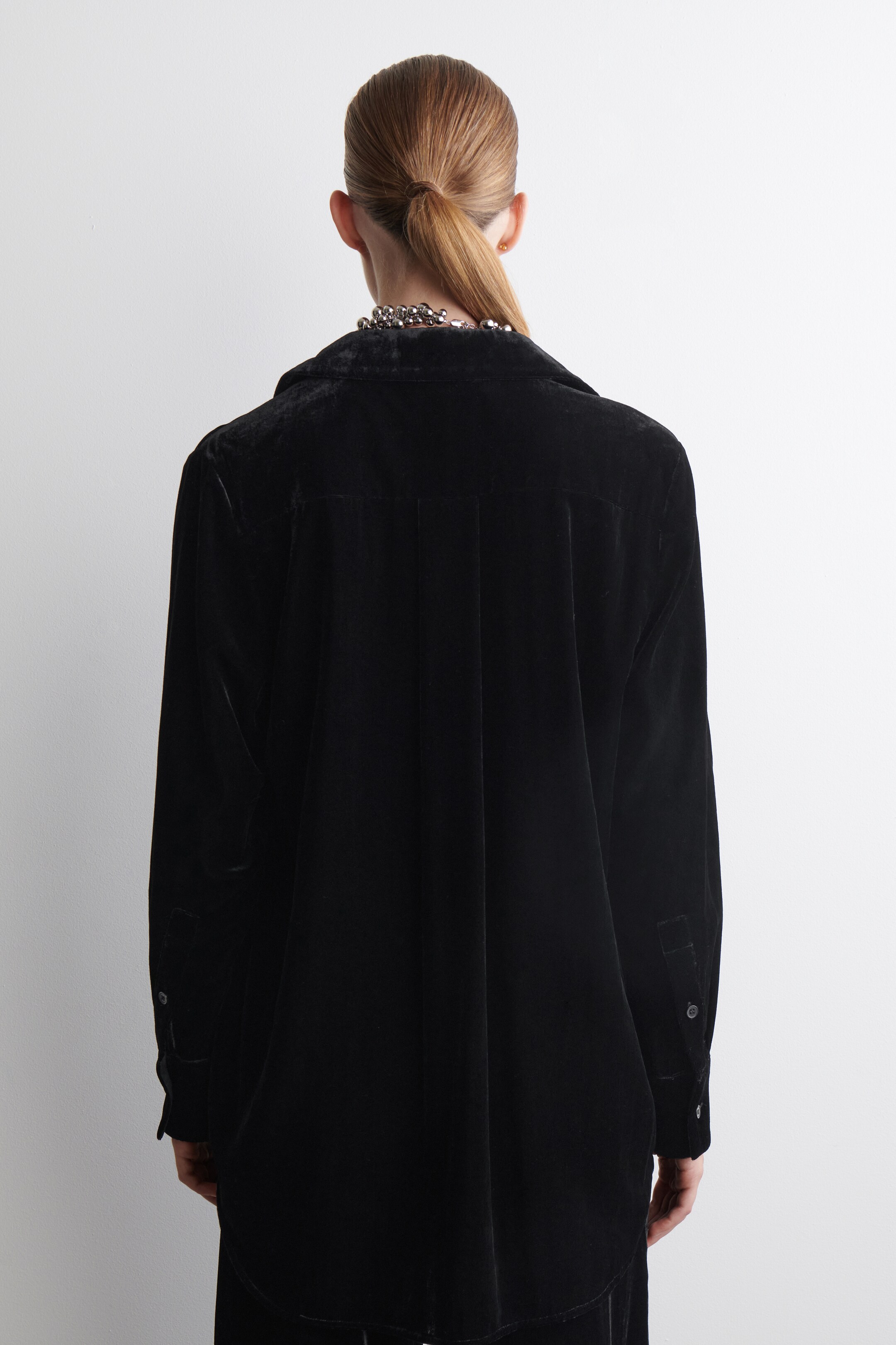 Visualizza immagine più grande: Una camicia di velluto nero viene mostrata da dietro, mostrando una vestibilità rilassata e oversize. Presenta un colletto classico, maniche lunghe con polsini abbottonati e pieghe verticali che dettagliamo la parte superiore della schiena.