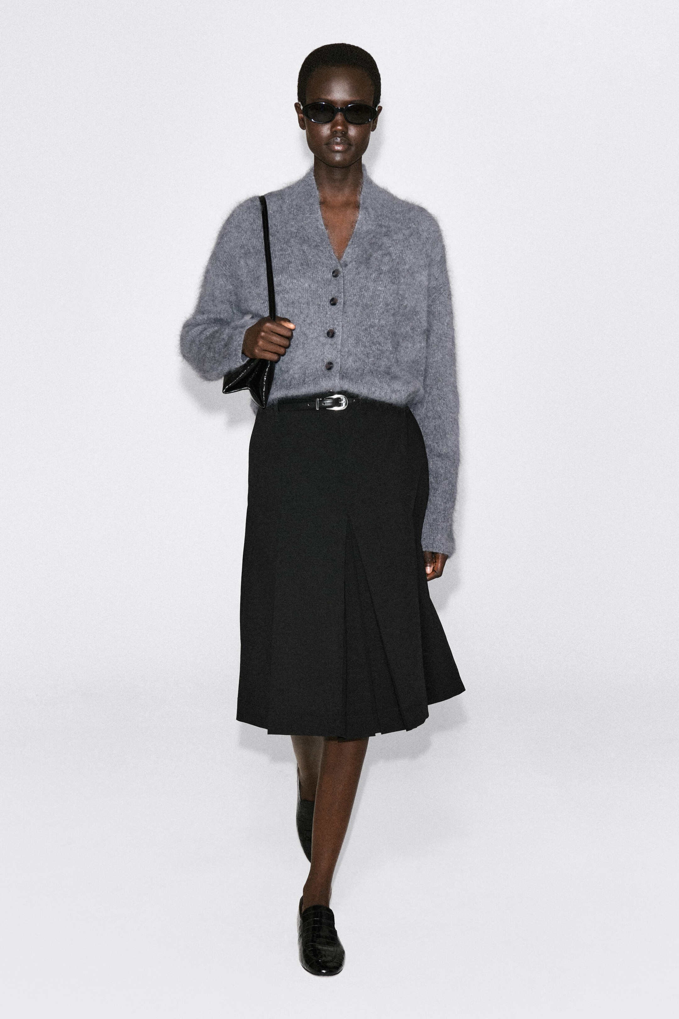 The draped brushed merino wool cardigan in gray is softly draped in a front view, featuring a fuzzy brushed texture, a V-neckline, and dark front buttons. It is styled tucked into a belted black skirt, showcasing its relaxed fit and cozy structure.