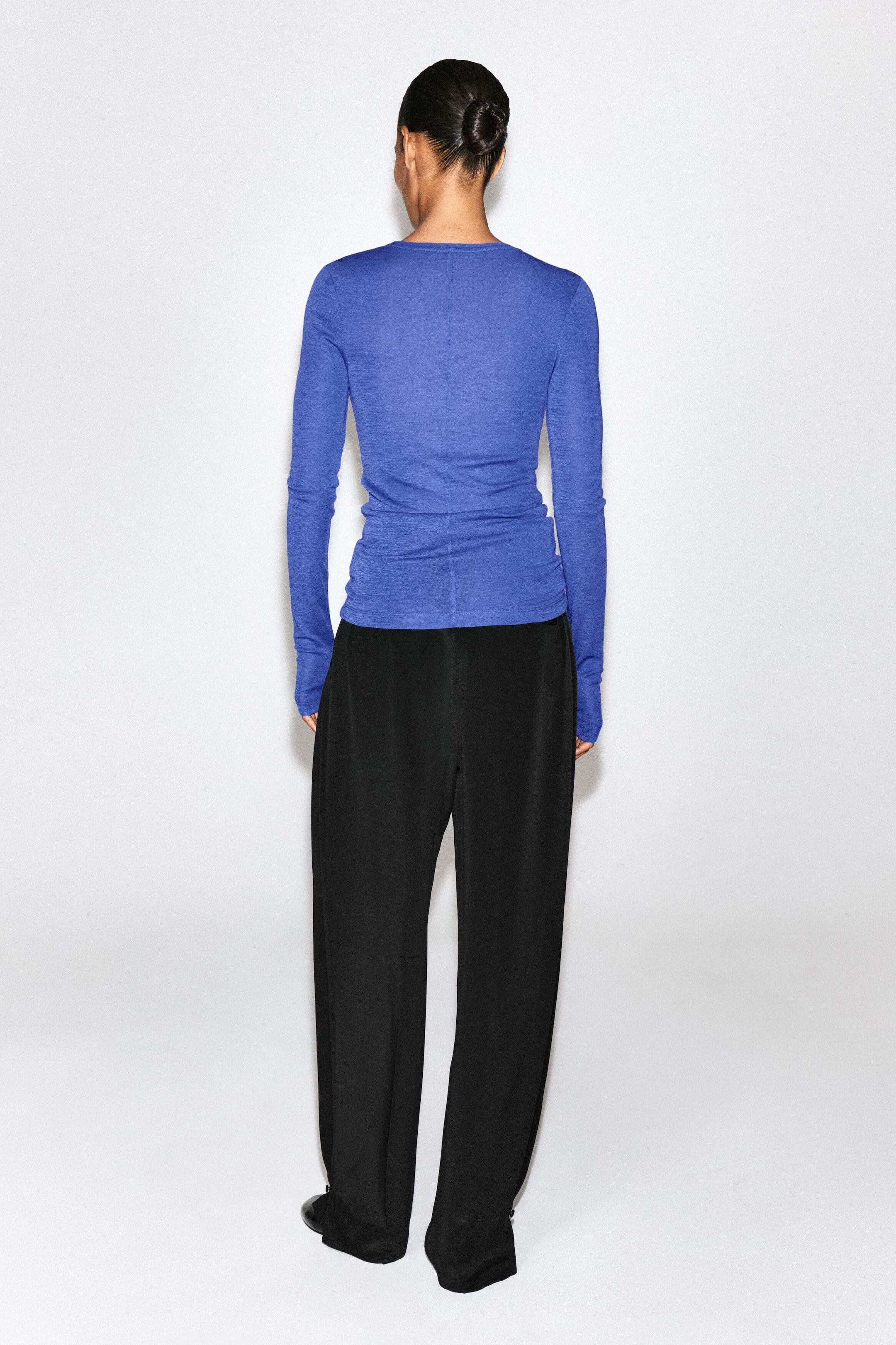 A blue slim-fit merino wool top with long sleeves and a visible centre-back seam is worn by the model, who is facing away from the camera. The top is styled with wide-leg black trousers. A blue slim-fit merino wool top with long sleeves and a visible centre-back seam is worn by the model, who is facing away from the camera. The top is styled with wide-leg black trousers.