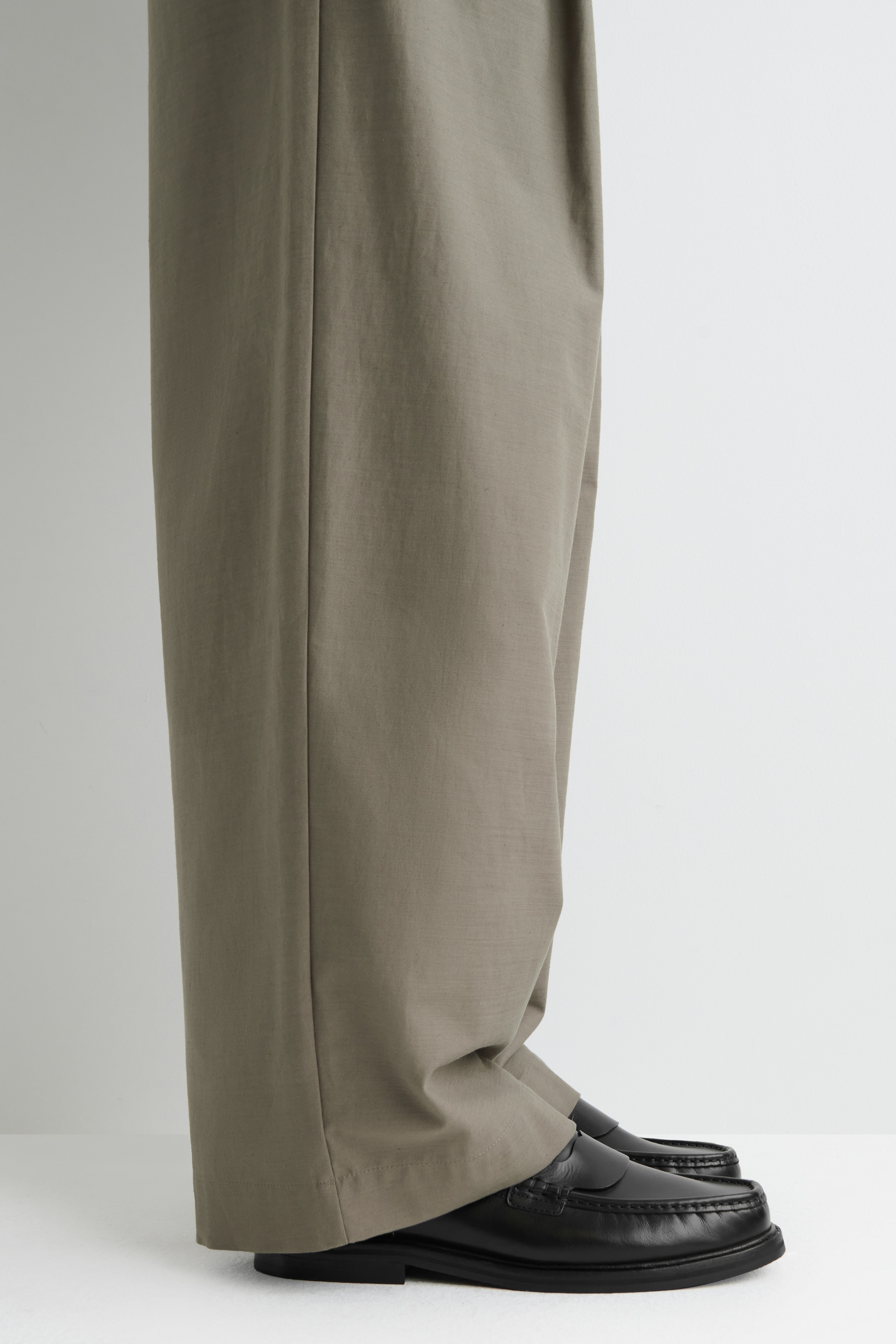 Visualizza immagine più grande: I pantaloni khaki a gamba larga presentano una piega centrale prominente e si accumulano sopra mocassini in pelle nera. I mocassini in pelle nera sono dettagliati con cuciture moc-toe e una suola spessa, come si vede da un profilo laterale.