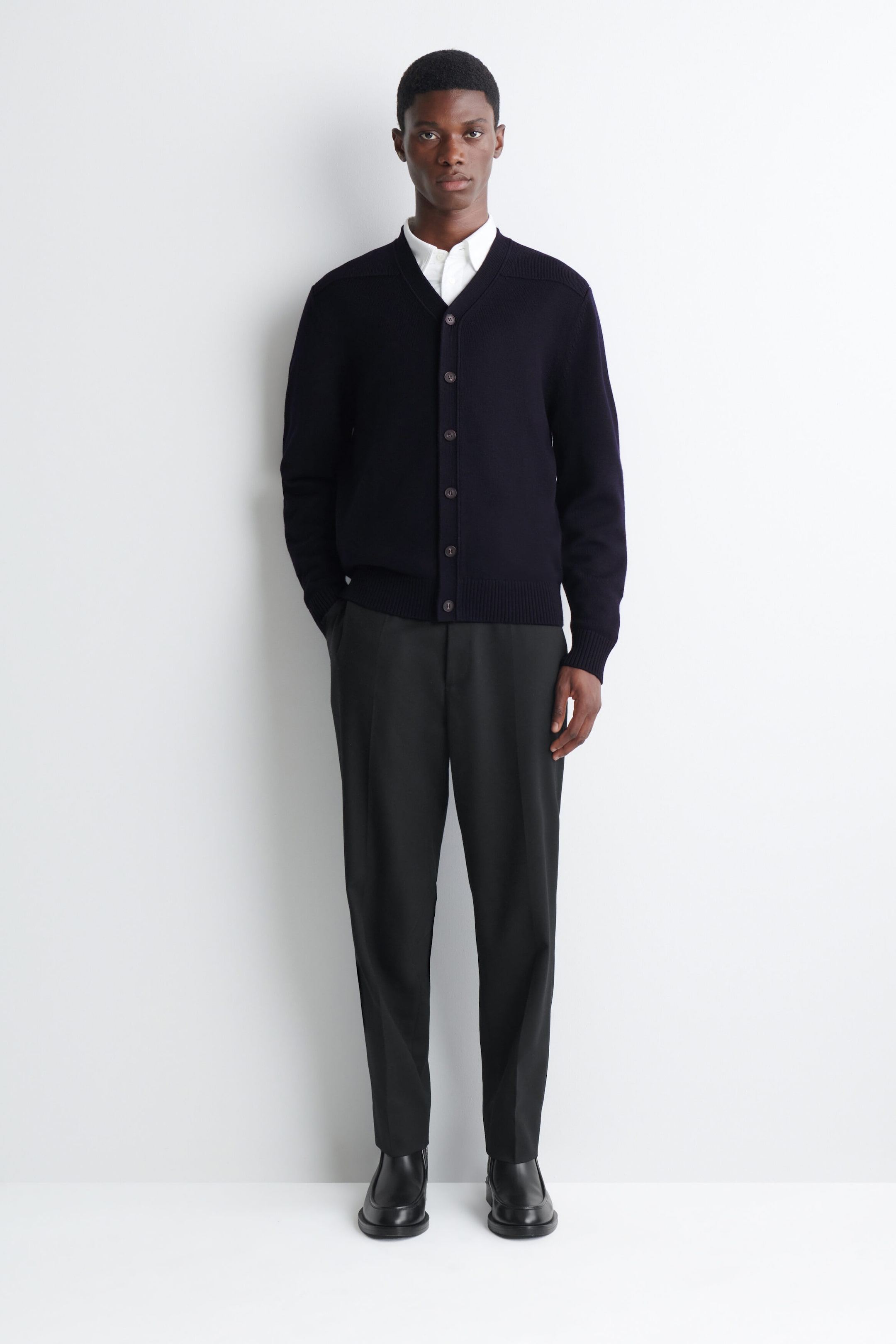 A dark navy-blue knit cardigan with a V-neckline and front button fastening is worn by the model over a white collared shirt. The cardigan features a ribbed hem and cuffs, and is styled with black pressed trousers and black leather shoes. A dark navy-blue knit cardigan with a V-neckline and front button fastening is worn by the model over a white collared shirt. The cardigan features a ribbed hem and cuffs, and is styled with black pressed trousers and black leather shoes.