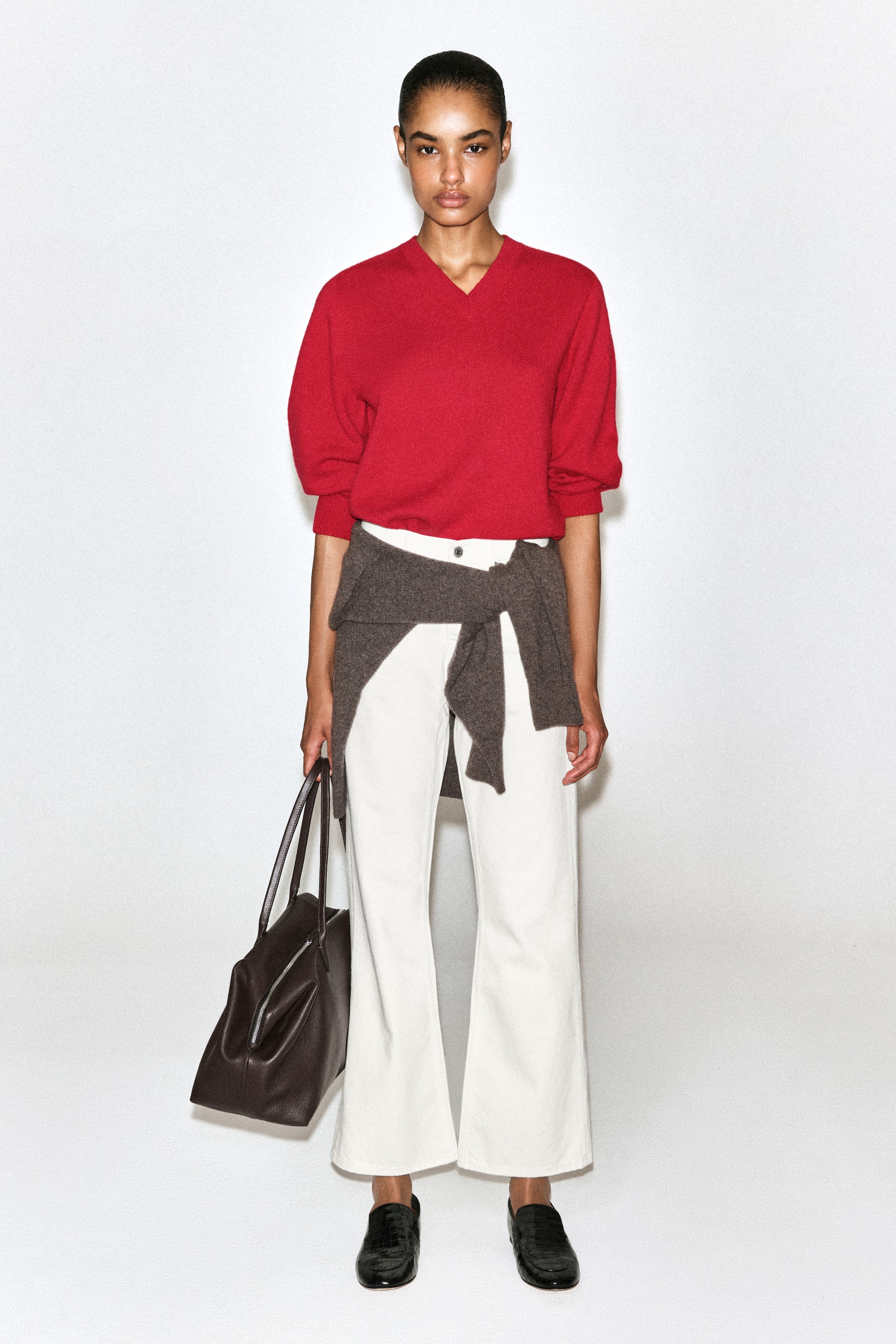 Ver imagen más grande: Los vaqueros bootcut en blanco roto se muestran de frente, con tiro alto y un ajuste entallado en los muslos que se abre sutilmente en el bajo recortado, a la altura del tobillo. Se combinan con un top rojo metido por dentro de la cintura y un jersey marrón anudado a la cadera.