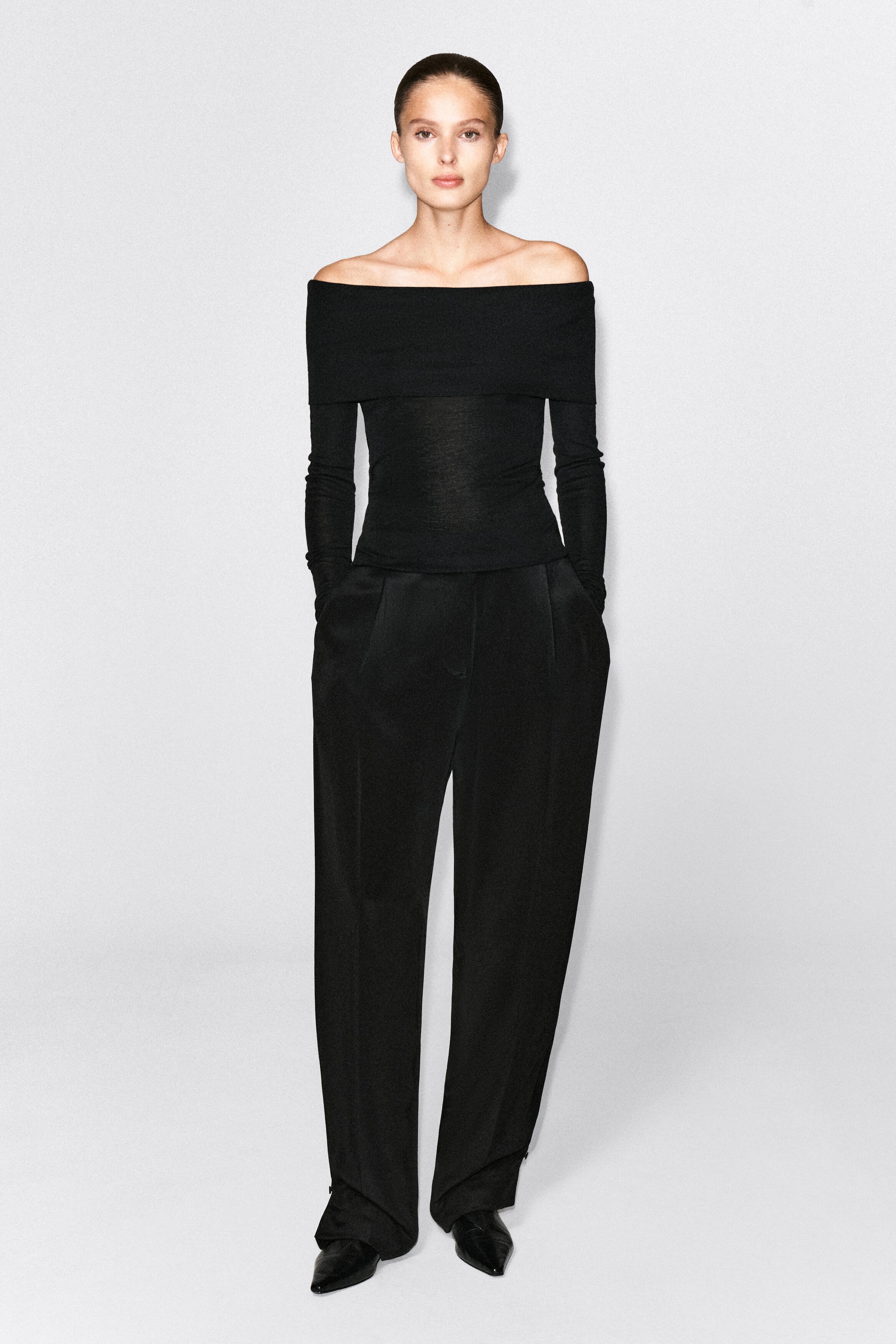 The model is wearing a black off-the-shoulder merino wool top featuring a wide fold-over neckline and long sleeves. This fitted, fine-knit top is styled with high-waisted, wide-leg black trousers.