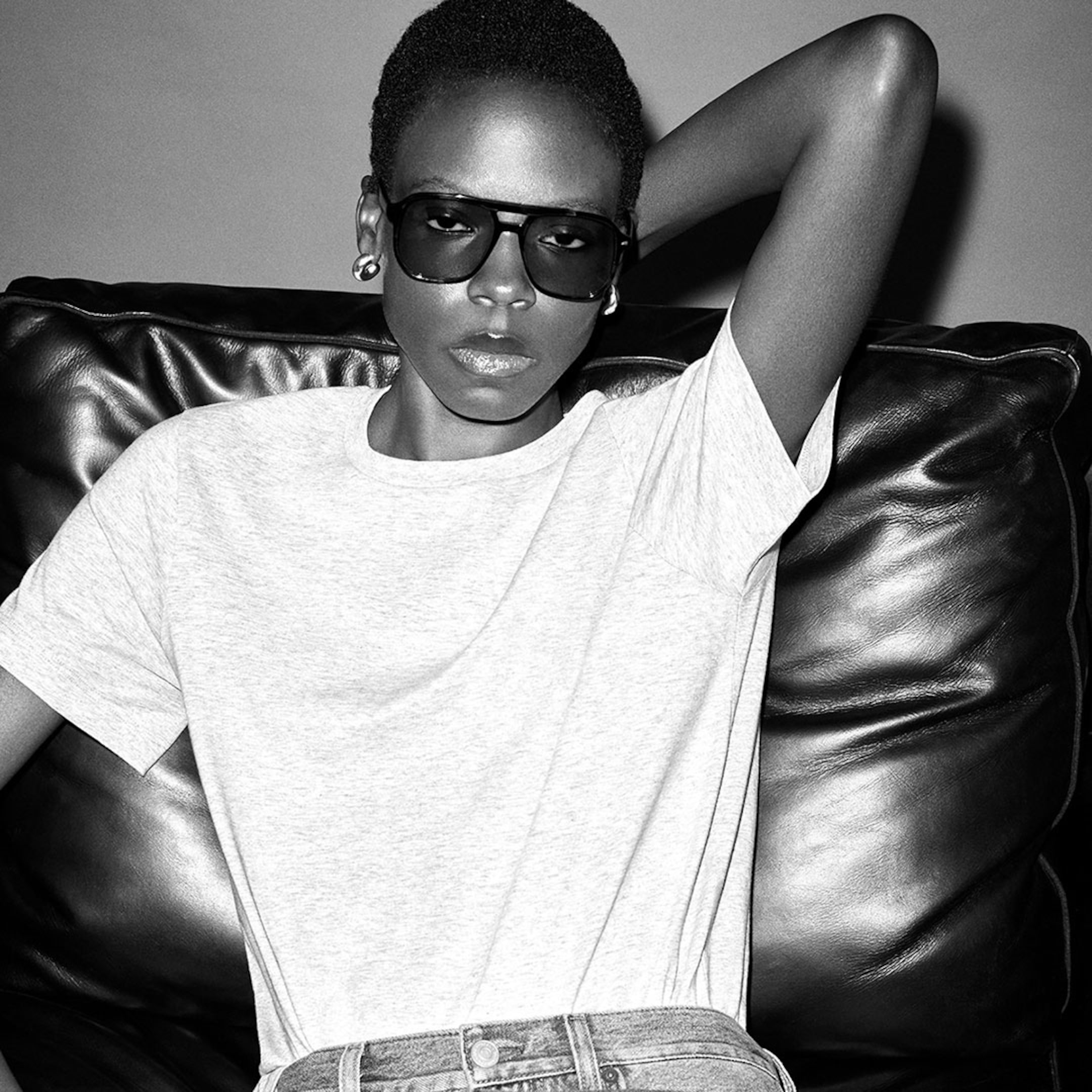 Person wearing sunglasses and a plain T-shirt, sitting on a leather couch, with one arm behind their head, in a relaxed pose. Black and white image.