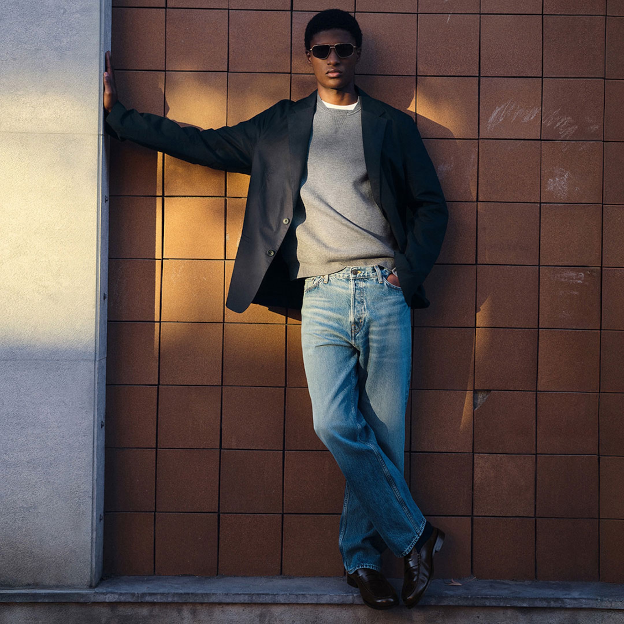 Person in sunglasses, dark blazer, gray sweater, and jeans leans against a tiled wall, casting a shadow in the sunlight.