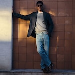 Placeholder for: Person in sunglasses, dark blazer, gray sweater, and jeans leans against a tiled wall, casting a shadow in the sunlight.