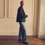 Placeholder for: Person in a dark jacket and jeans stands indoors against a neutral-toned wall, looking sideways.