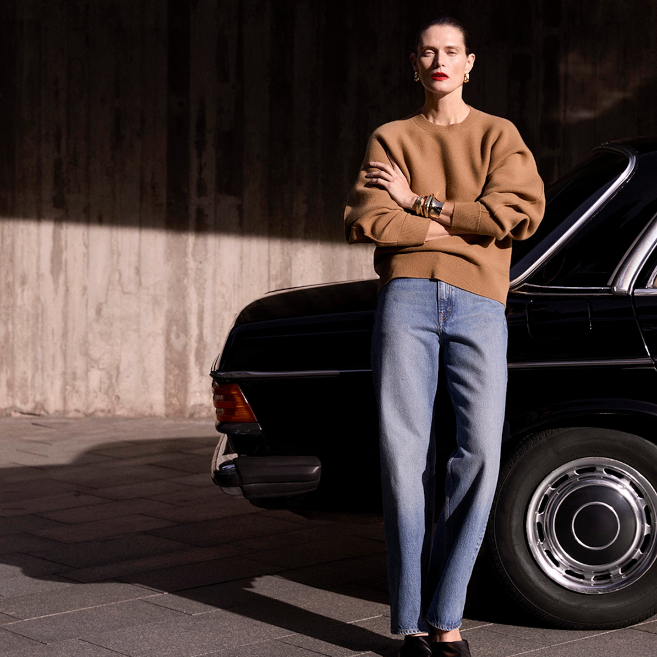 Woman in a brown sweater and jeans stands confidently with arms crossed beside a black car against a textured wall.