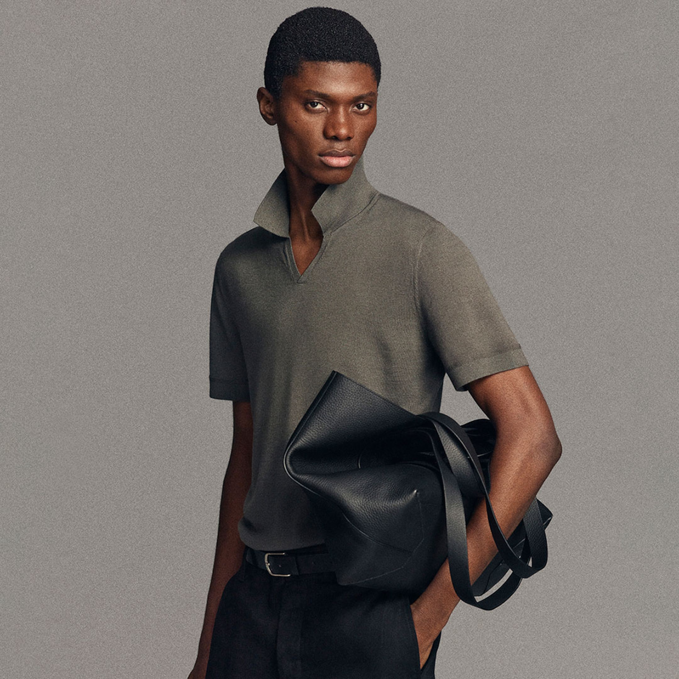 Model in a gray shirt with a turned-up collar holds a large black leather bag against a neutral background.