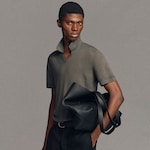 Placeholder for: Model in a gray shirt with a turned-up collar holds a large black leather bag against a neutral background.