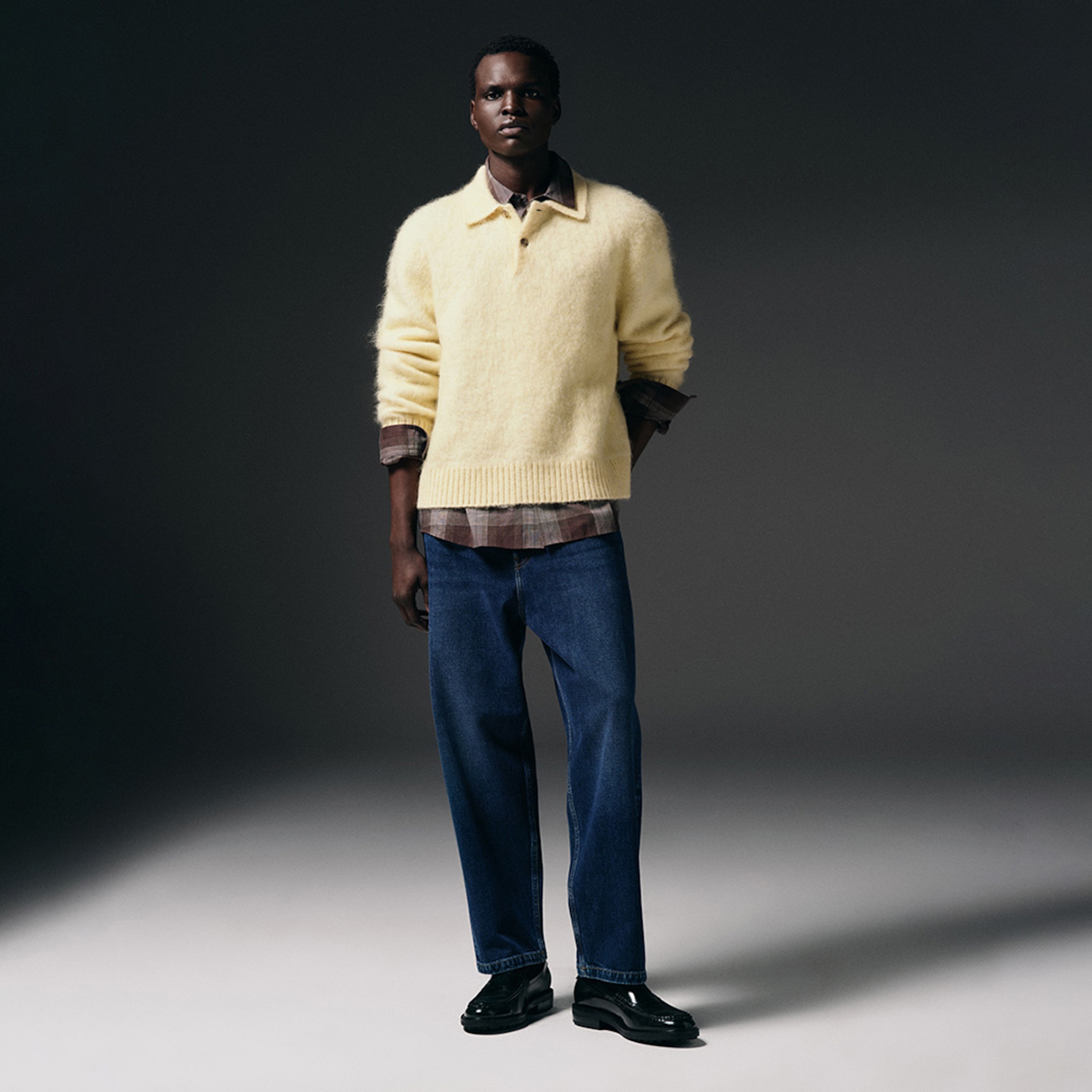 Person wearing a yellow fuzzy sweater over a plaid shirt, blue jeans, and black loafers, posing against a dark background.