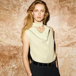 Placeholder for: A person with long hair wearing a sleeveless beige top with a unique neckline and black pants, standing against a marbled background.
