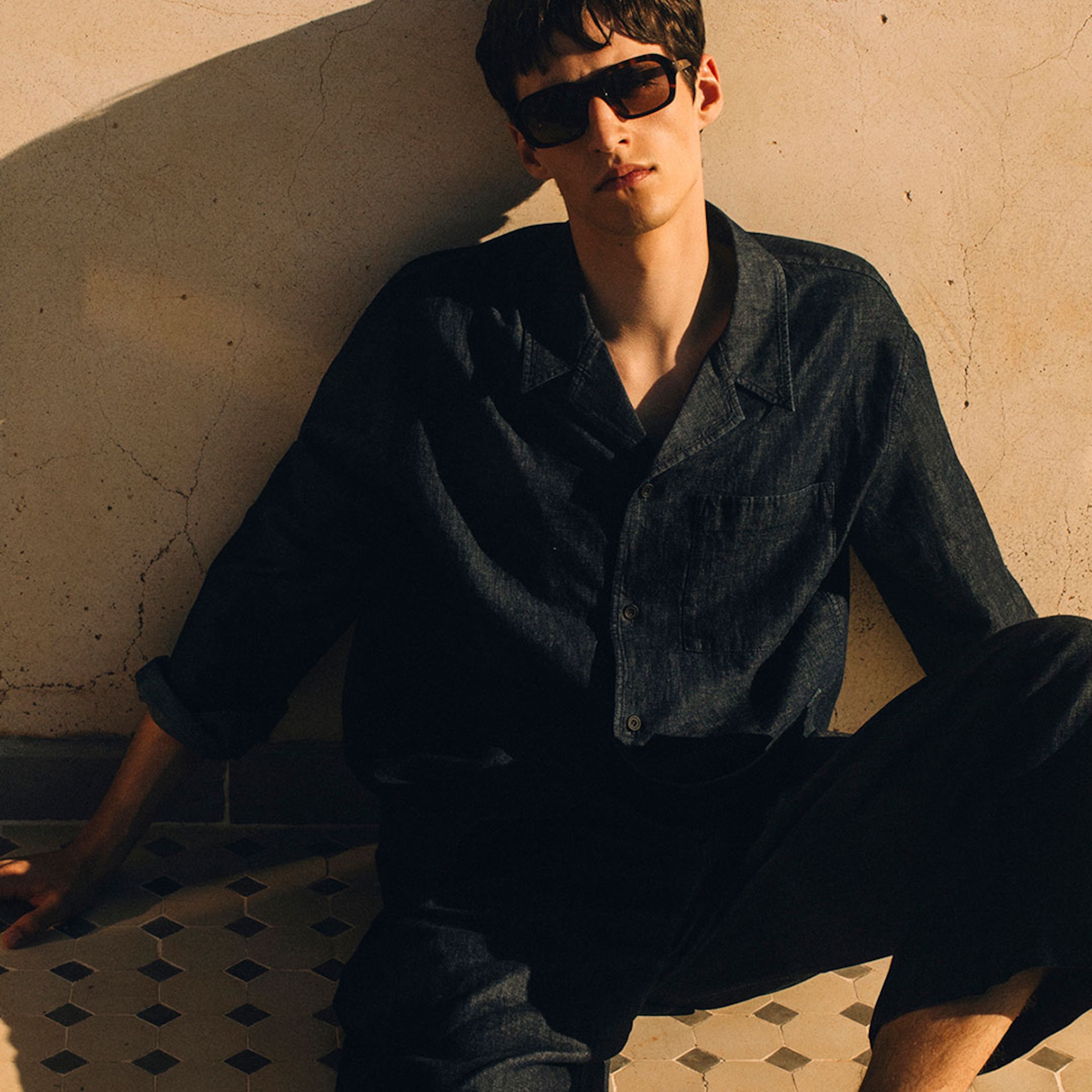 A sunlit lifestyle shot of a man seated on a patterned tiled floor, leaning against a textured cream wall. He is wearing a dark-wash linen long-sleeved shirt with tonal buttons and a classic collar, paired with matching dark linen trousers. He is styled with chunky dark sunglasses, and the warm, high-contrast shadows emphasize the heavy, premium texture of the linen fabric.