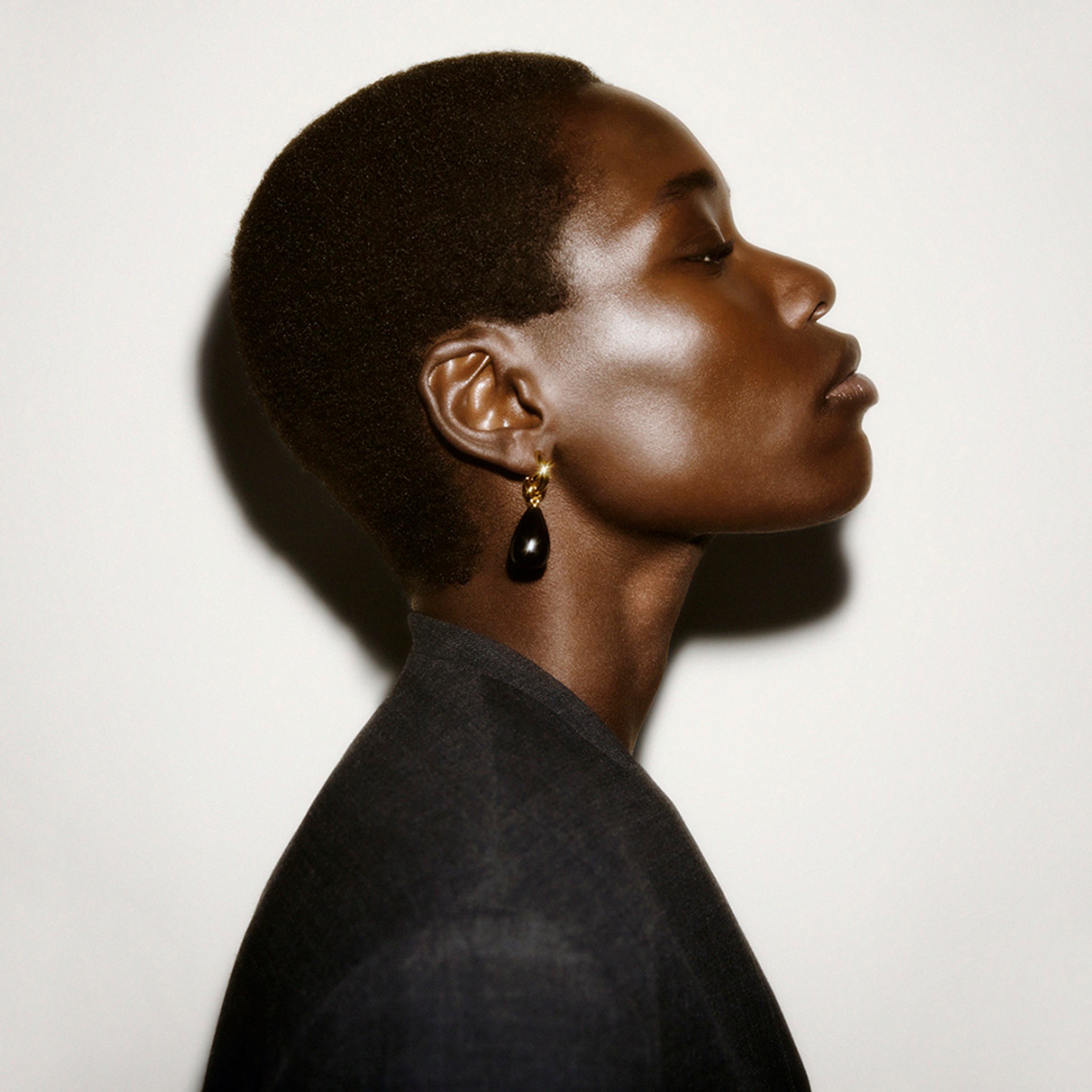Profile of a person with short hair wearing a black teardrop earring and a dark jacket against a light background.