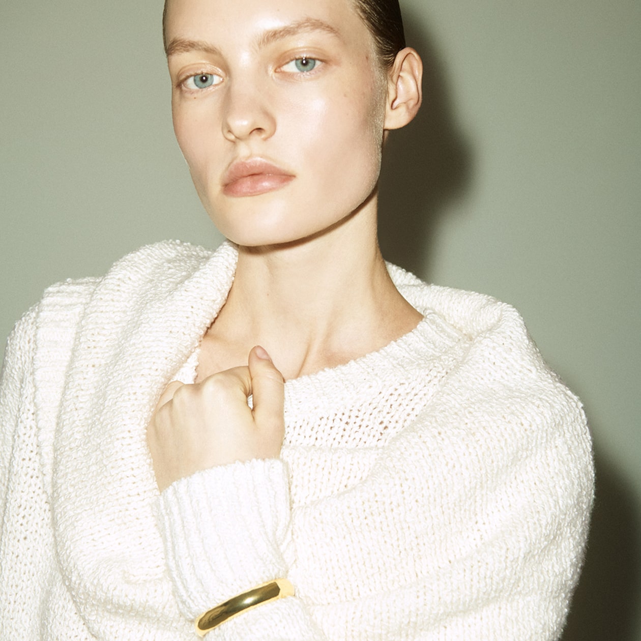 Person with slicked-back hair wearing a cozy white sweater and a gold bracelet, gazing directly at the camera.