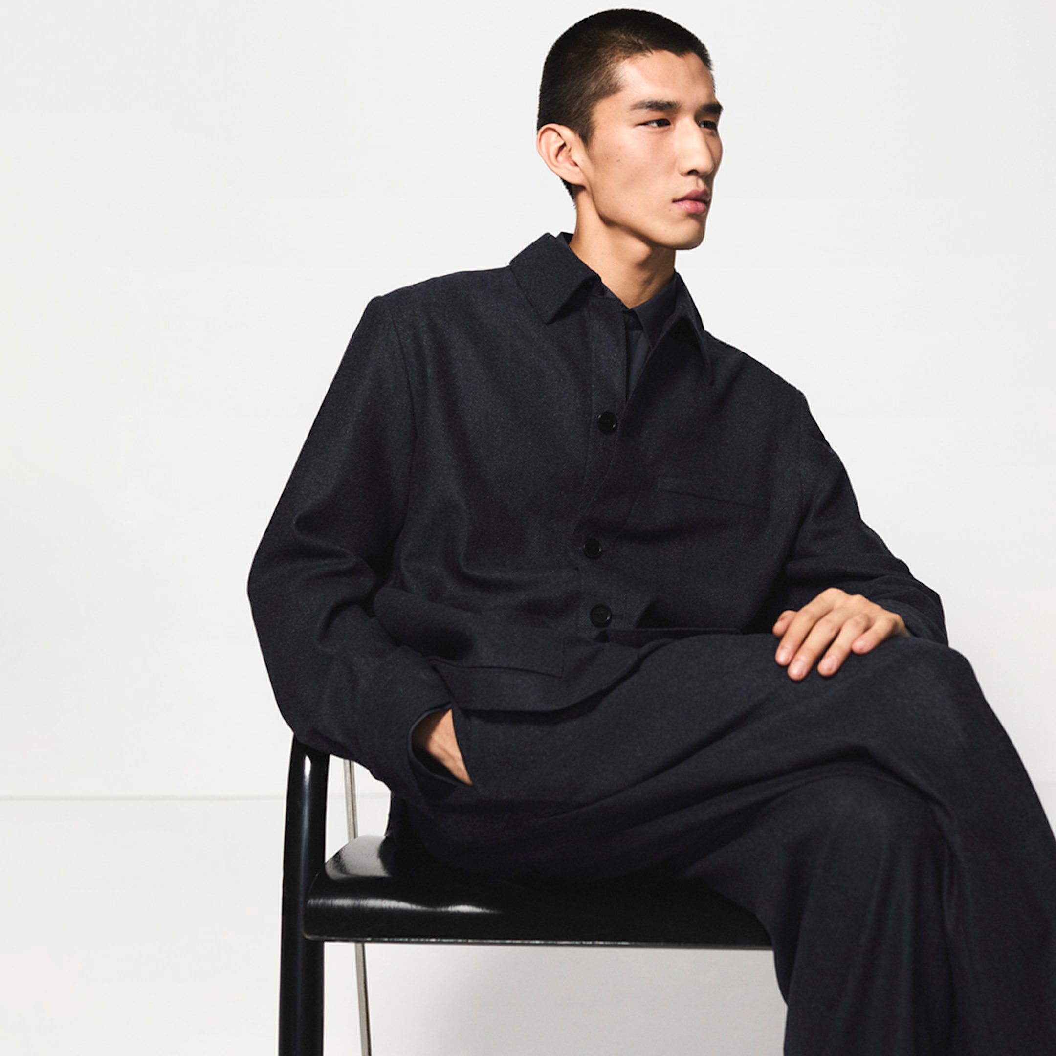 Person in black button-up jacket and pants, sitting thoughtfully against a plain background with hands relaxed in lap.