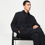 Placeholder for: Person in black button-up jacket and pants, sitting thoughtfully against a plain background with hands relaxed in lap.