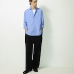 Placeholder for: Man in a blue shirt and black trousers stands against a white background, hands in pockets, looking to the side.