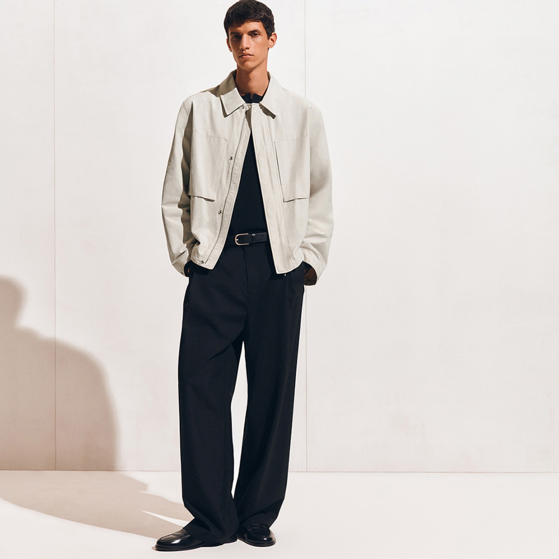 A person wearing a light beige jacket over a black shirt, with black wide-leg pants and black shoes, standing against a plain background.