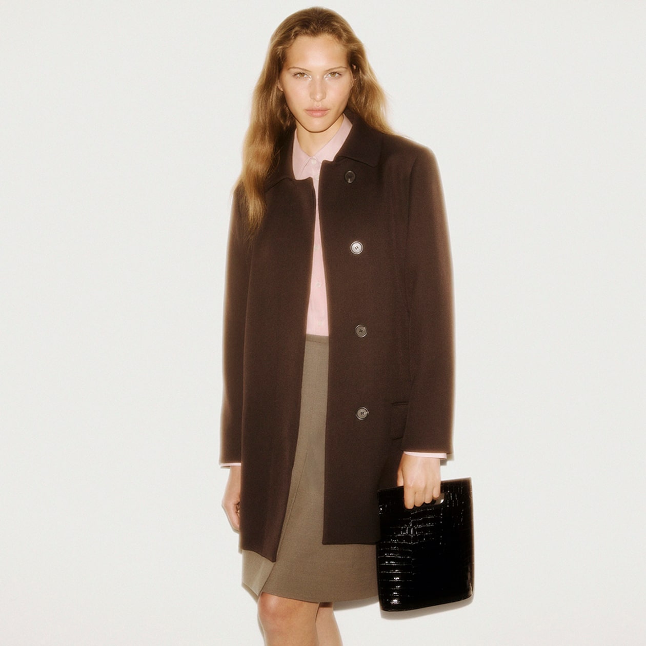 A person wearing a dark coat over a light pink shirt and beige skirt, holding a black textured clutch, poses against a neutral background.