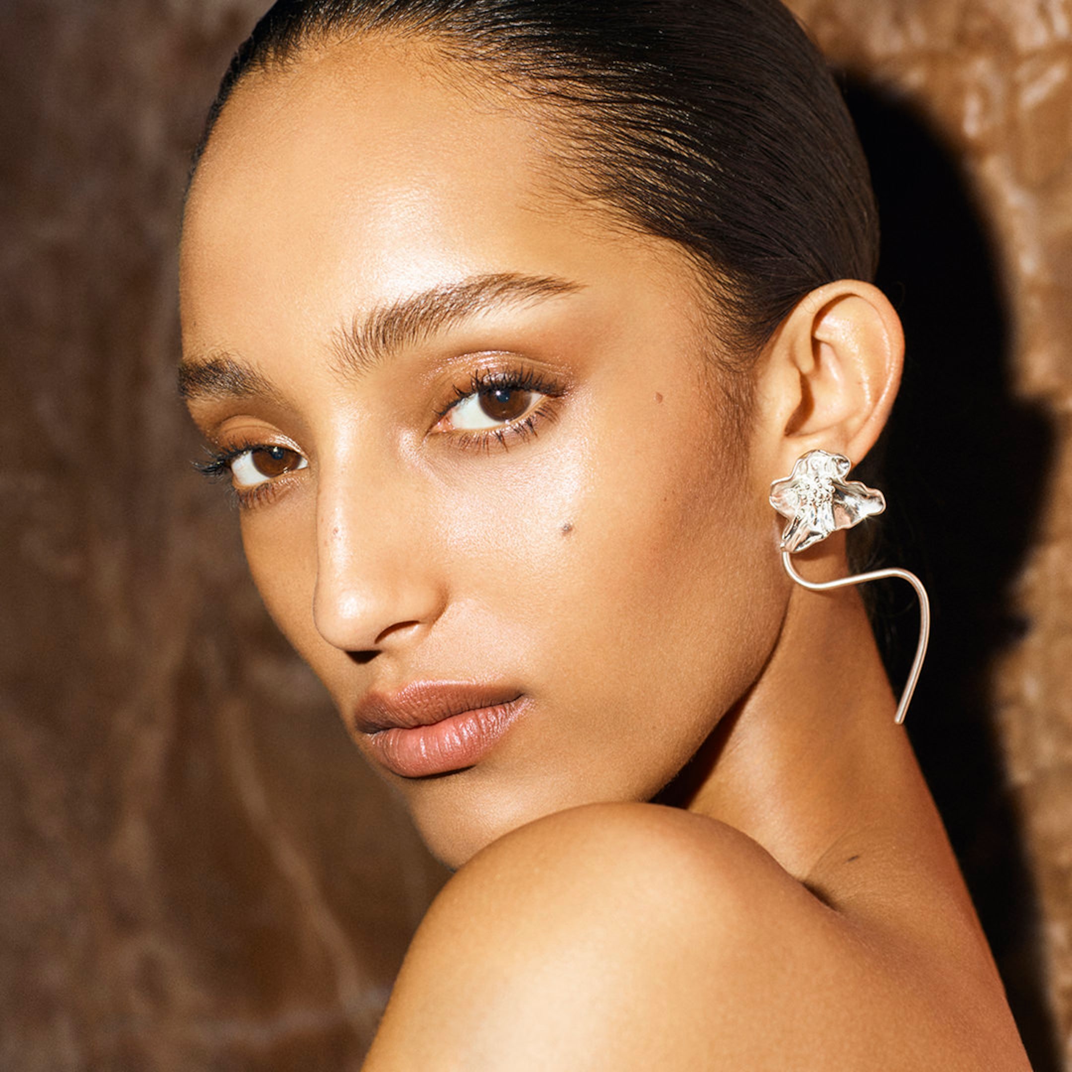 A person with sleek hair poses against a brown background, wearing a large, artistic earring shaped like a flower.