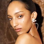 Placeholder for: A person with sleek hair poses against a brown background, wearing a large, artistic earring shaped like a flower.
