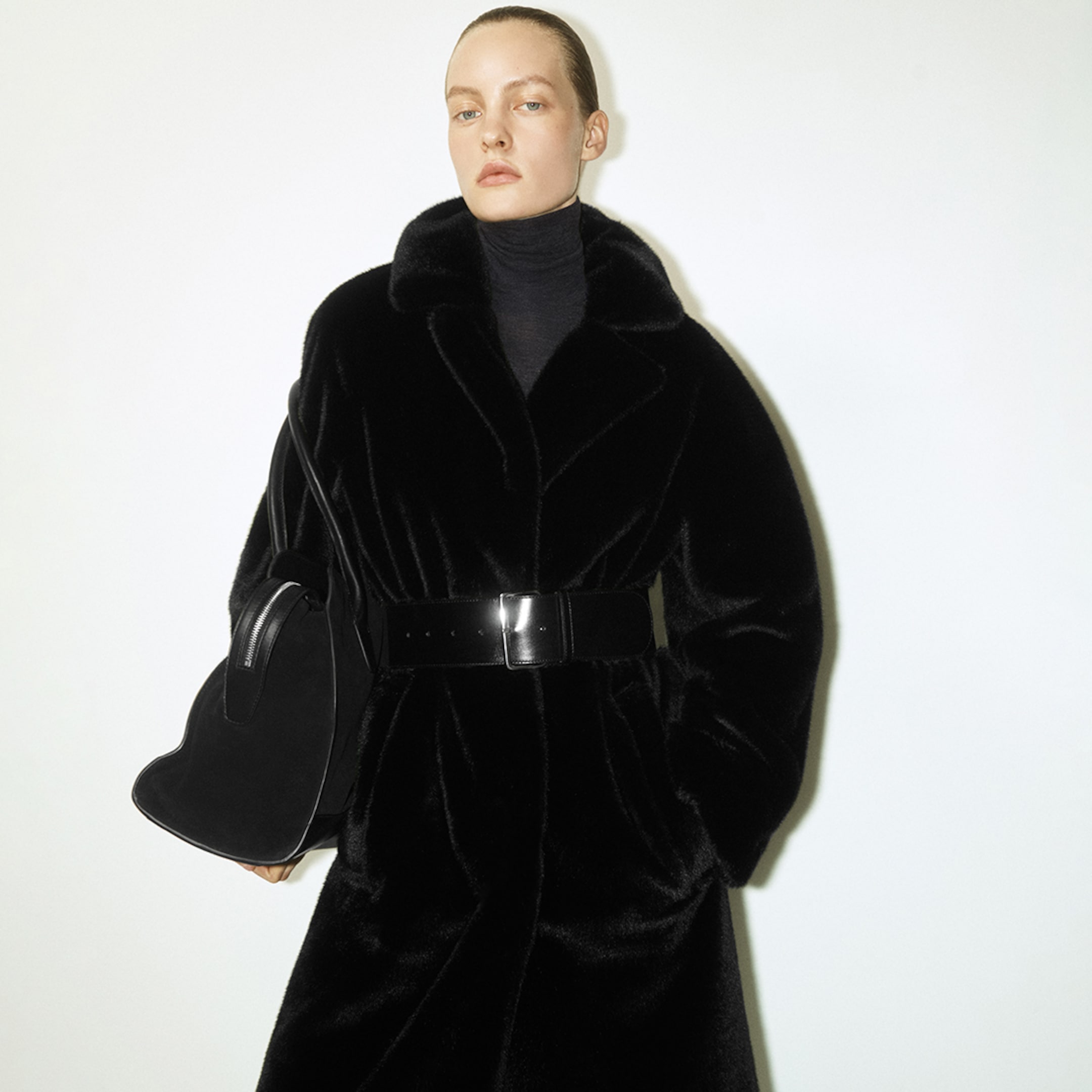 Person in a black fur coat with a wide belt, holding a matching bag, poses against a plain white background.