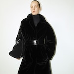 Placeholder for: Person in a black fur coat with a wide belt, holding a matching bag, poses against a plain white background.