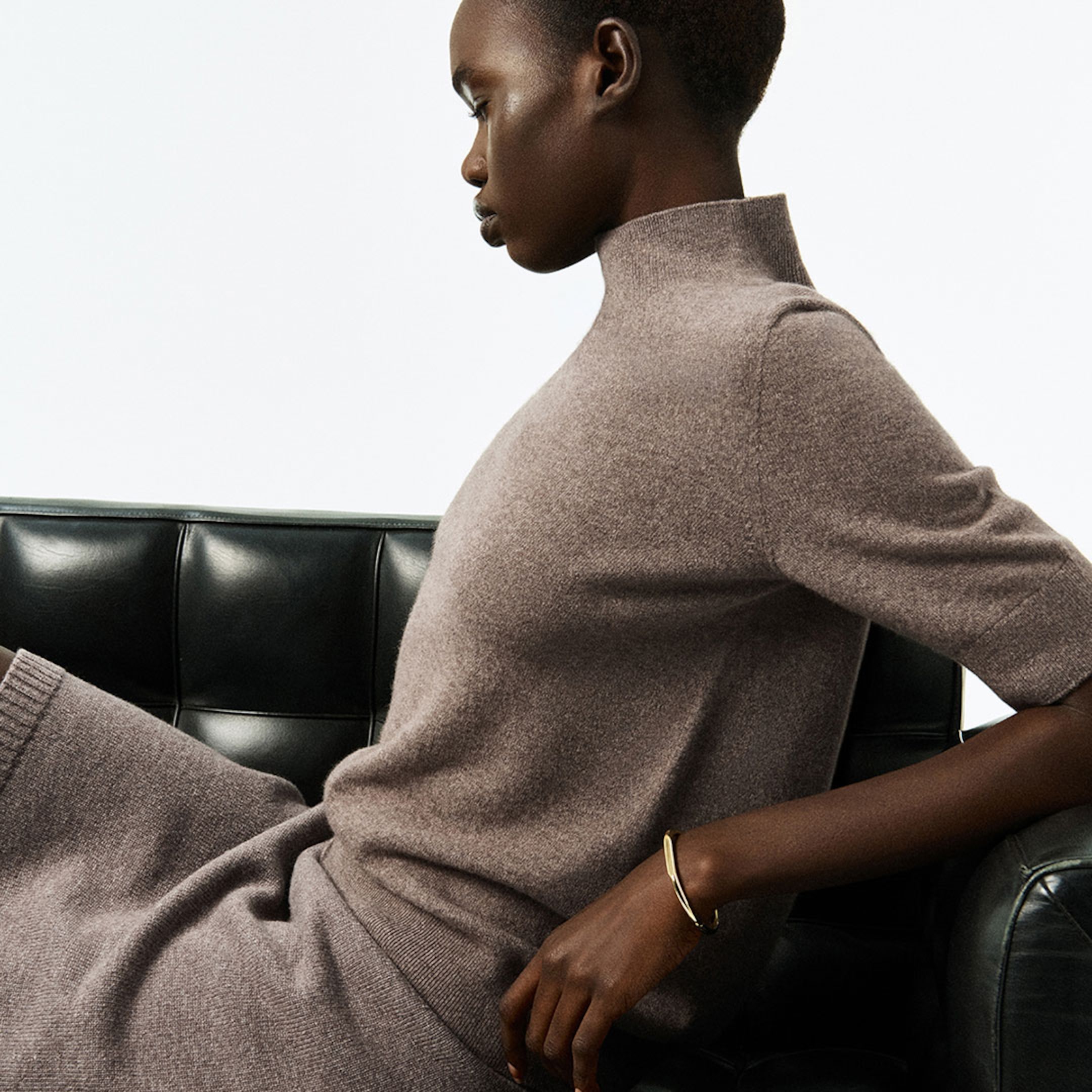 Model in a taupe turtleneck sweater dress, sitting on a black leather couch, looking off to the side.
