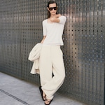 Placeholder for: A person wearing oversized cream pants, a white top, and sunglasses, leaning against a textured metal wall while holding a cream jacket.