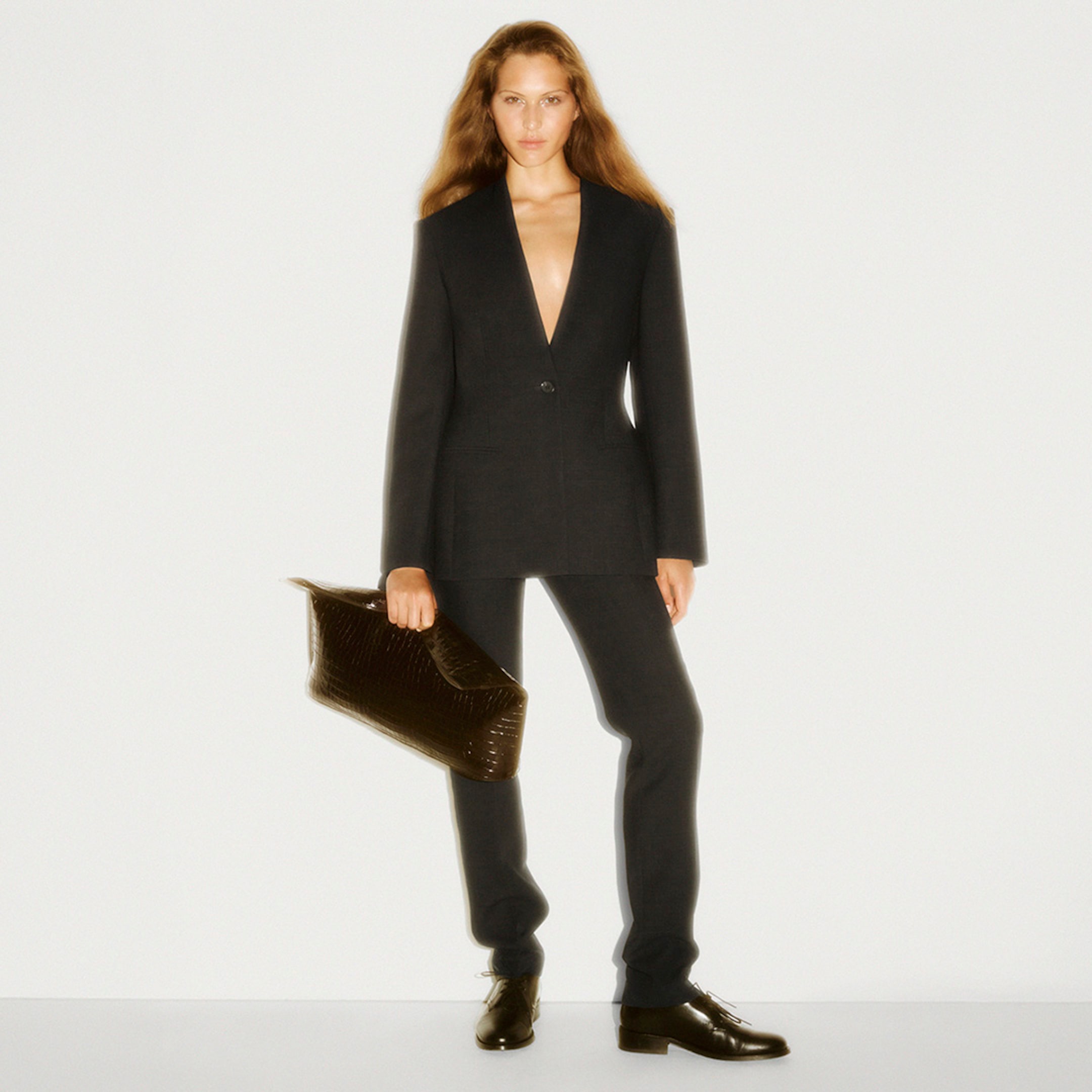 Woman in a black suit with a deep neckline holds a textured bag, standing against a plain backdrop.