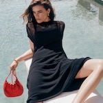 Placeholder for: Person in black dress lounging by a pool, holding a small red bag, with wavy brown hair and gold earrings. The background features clear water.