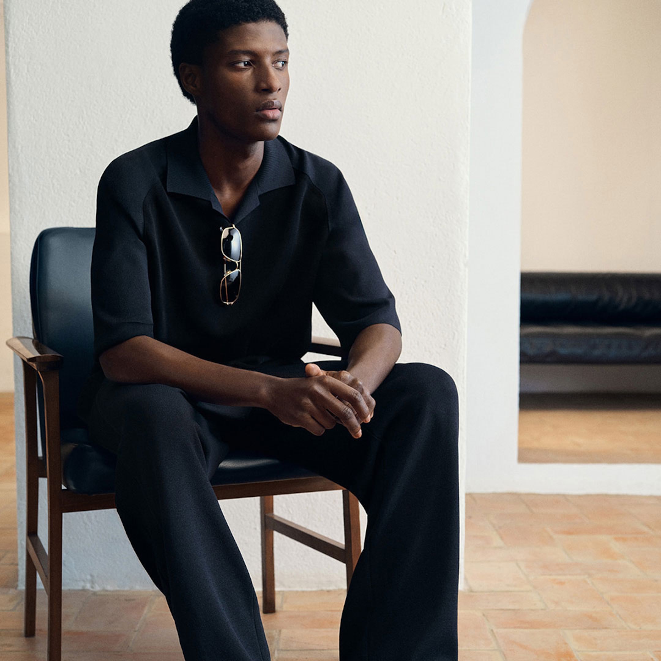 Man in a black outfit sits on a chair indoors, sunglasses hanging from his shirt, with a neutral expression.